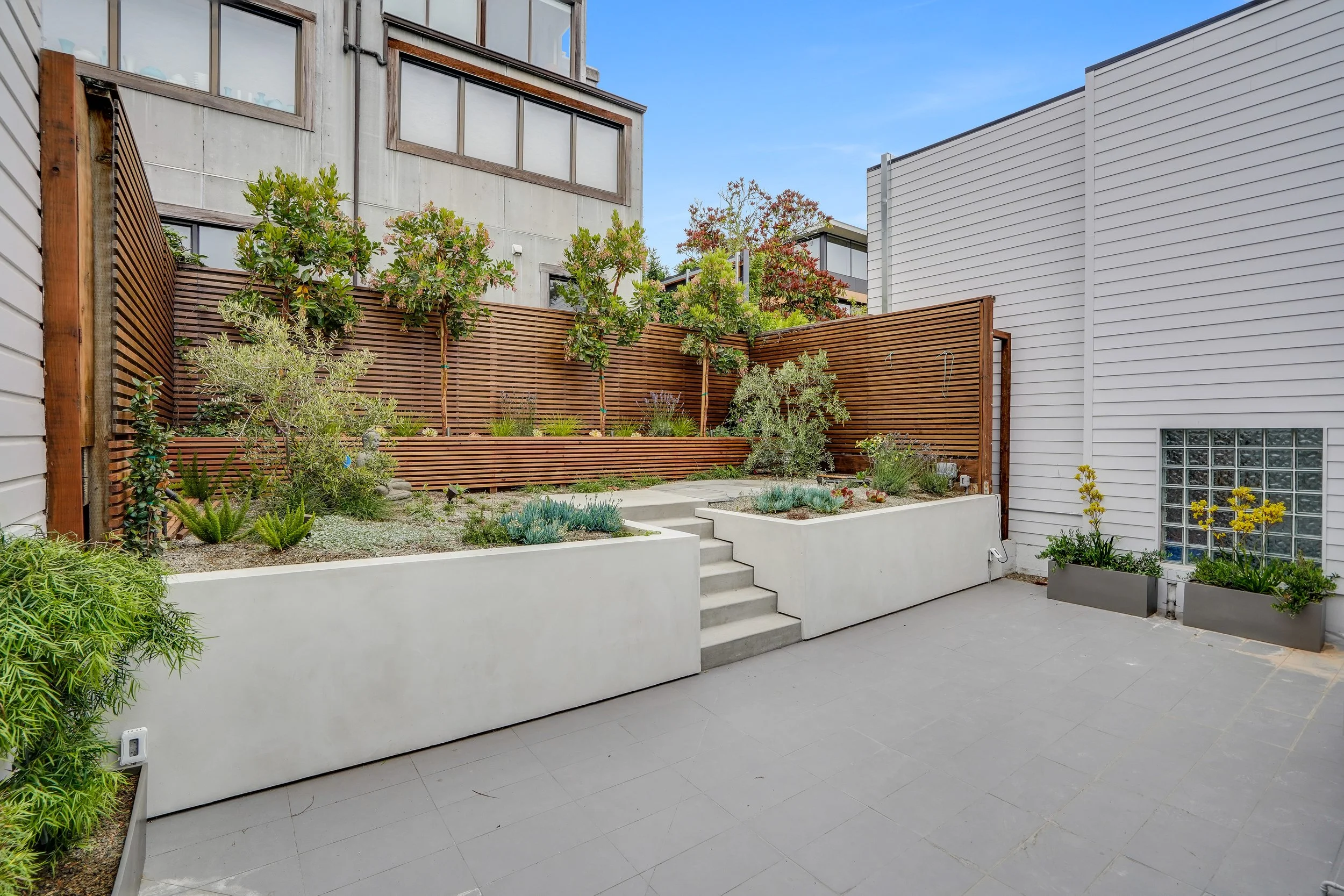 Backyard courtyard with a concrete retaining wall, steps, wood-slat fence, and landscaped planting beds