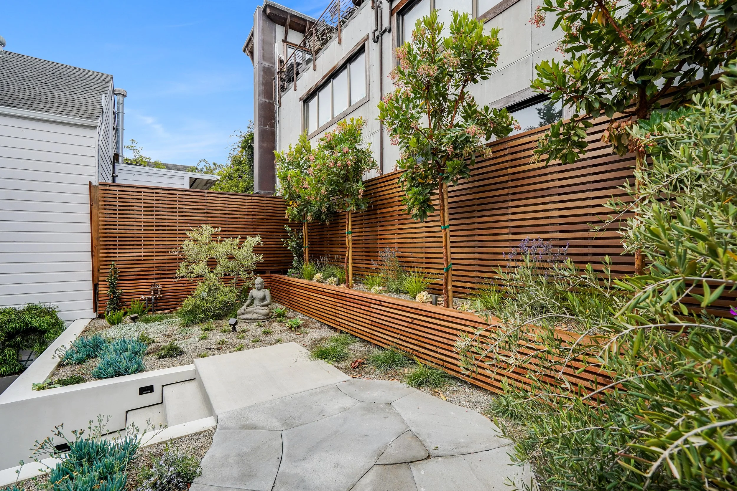 Backyard courtyard with a stone-paved patio, raised concrete planters, wood-slat privacy fence, and mature shrubs