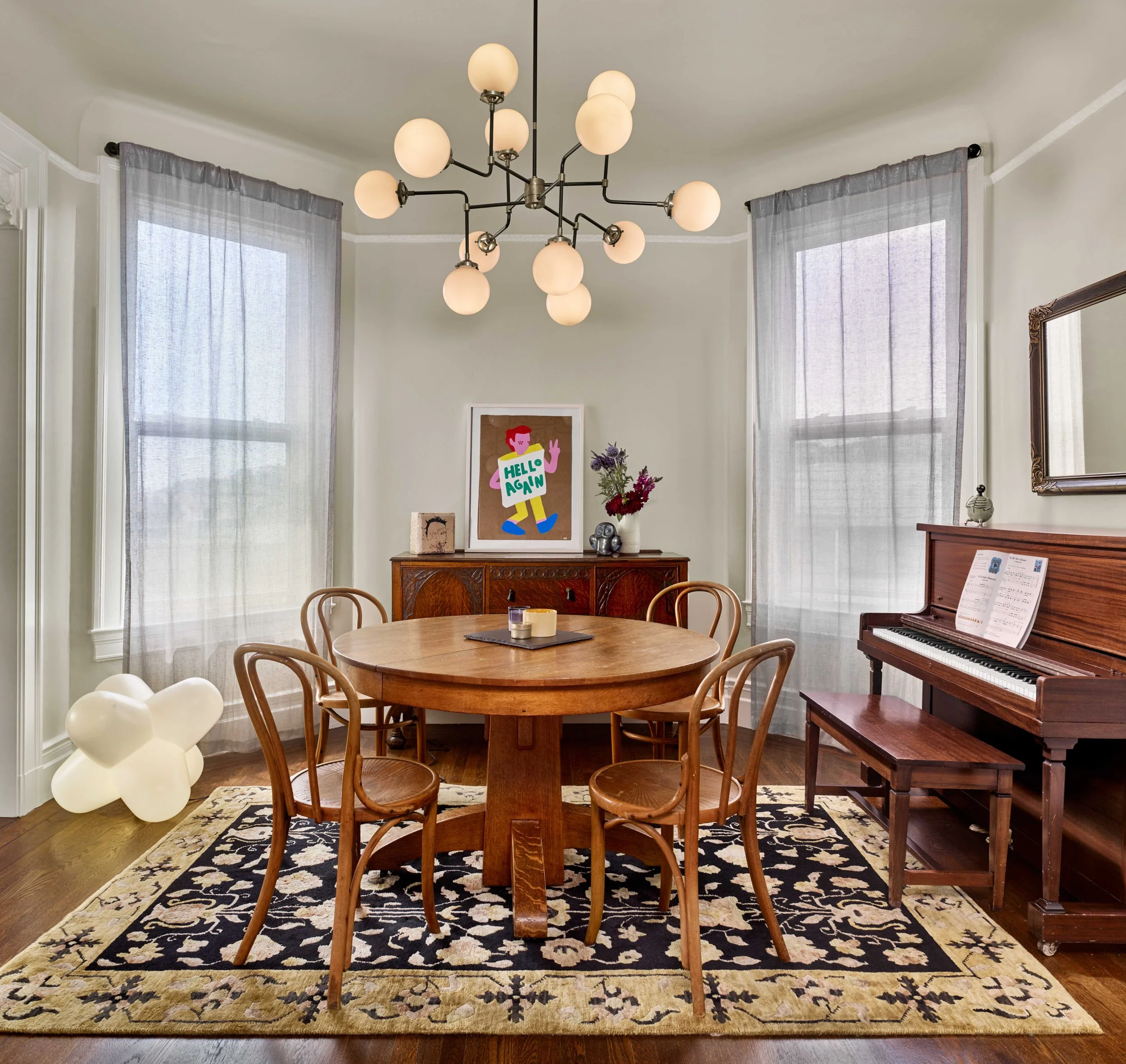 Dining room with a round wood table, modern globe chandelier, upright piano, patterned rug, and bay windows