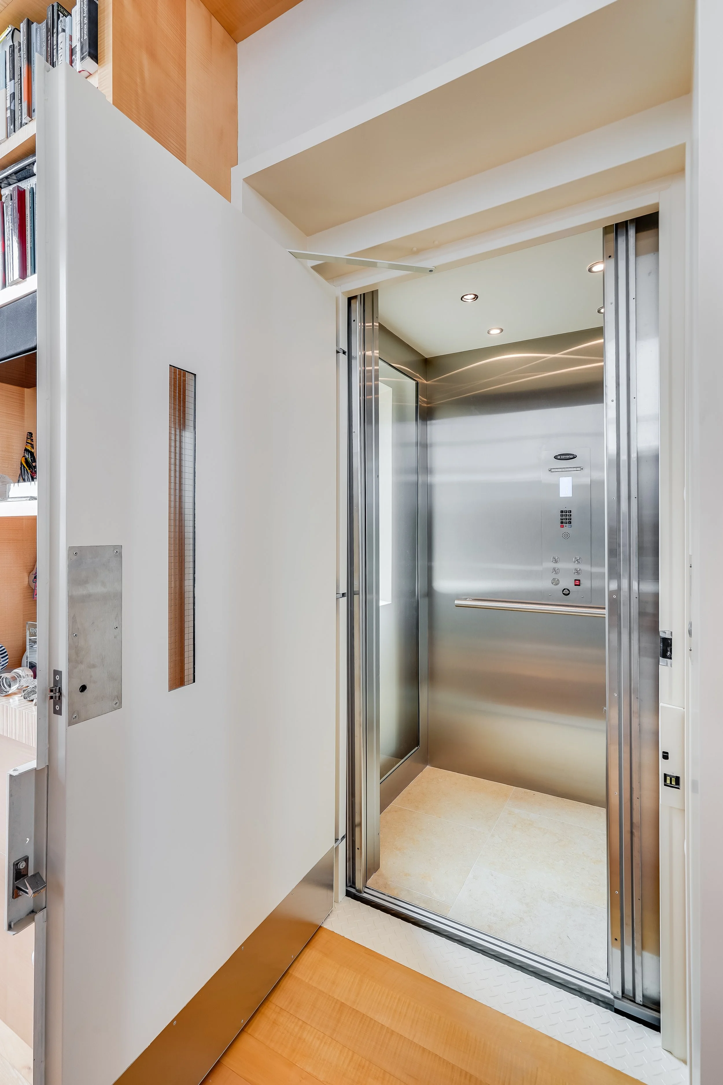 Stainless steel residential elevator with open doors, set into a hallway beside built-in shelving