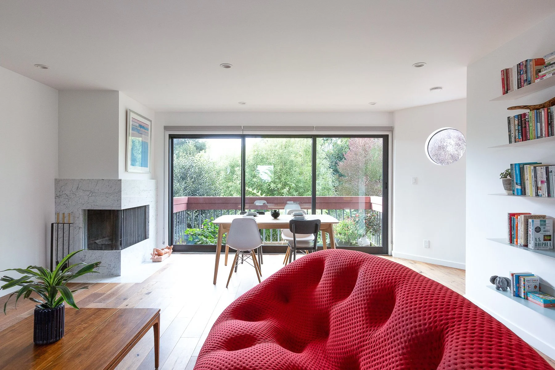 Living room with a stone fireplace, red tufted lounge chair, built-in bookshelves, and a dining area by large windows