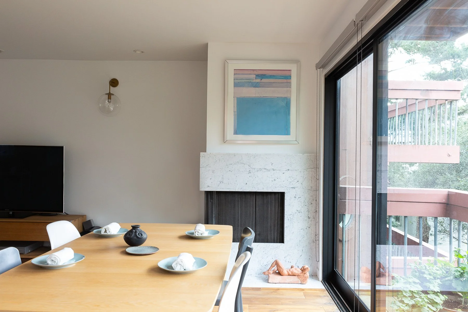 Dining area with a light wood table set for tea, a marble fireplace, and floor-to-ceiling windows overlooking greenery