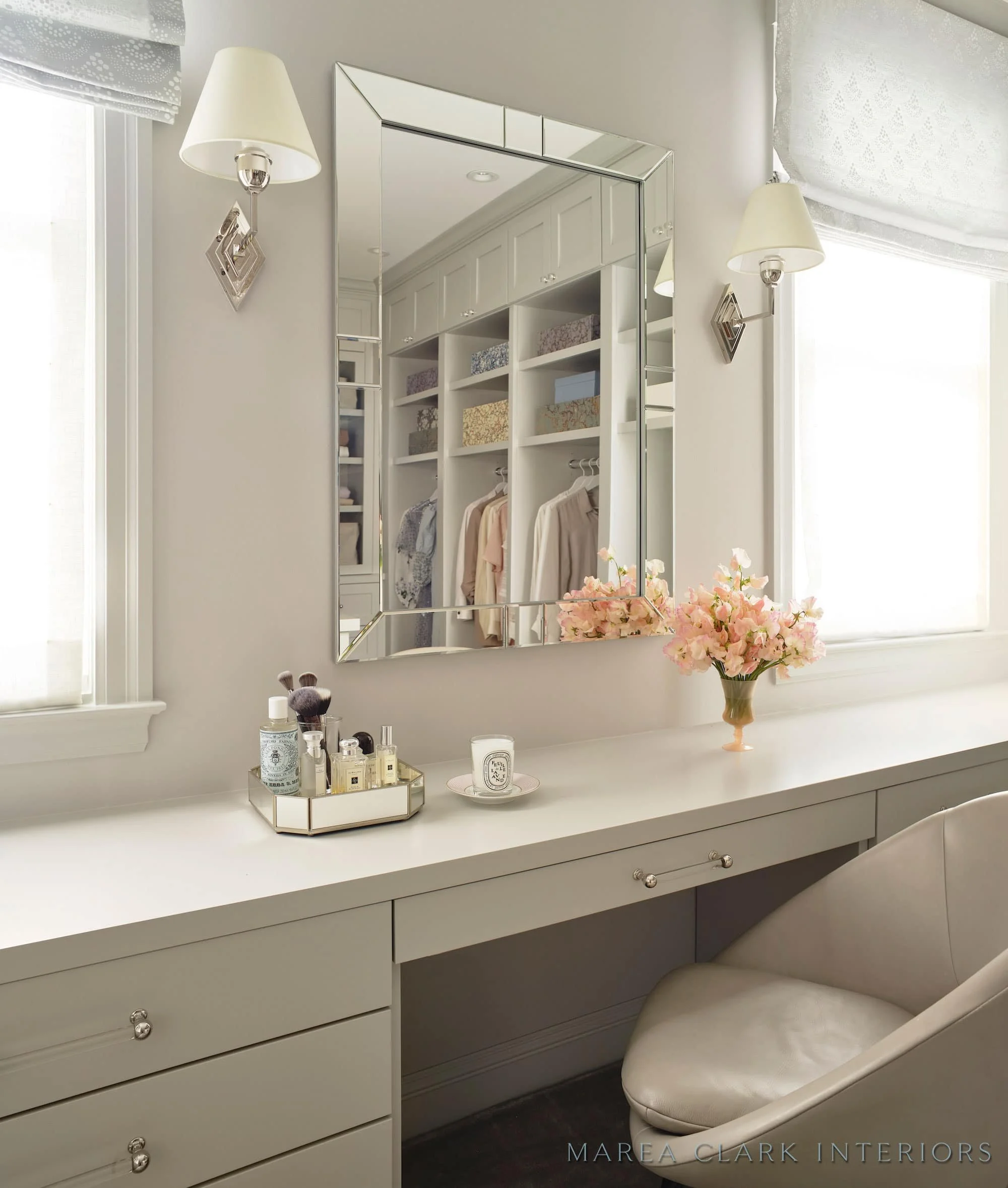 Dressing vanity with a mirror, wall sconces, an upholstered stool, and glass-front cabinets by a window