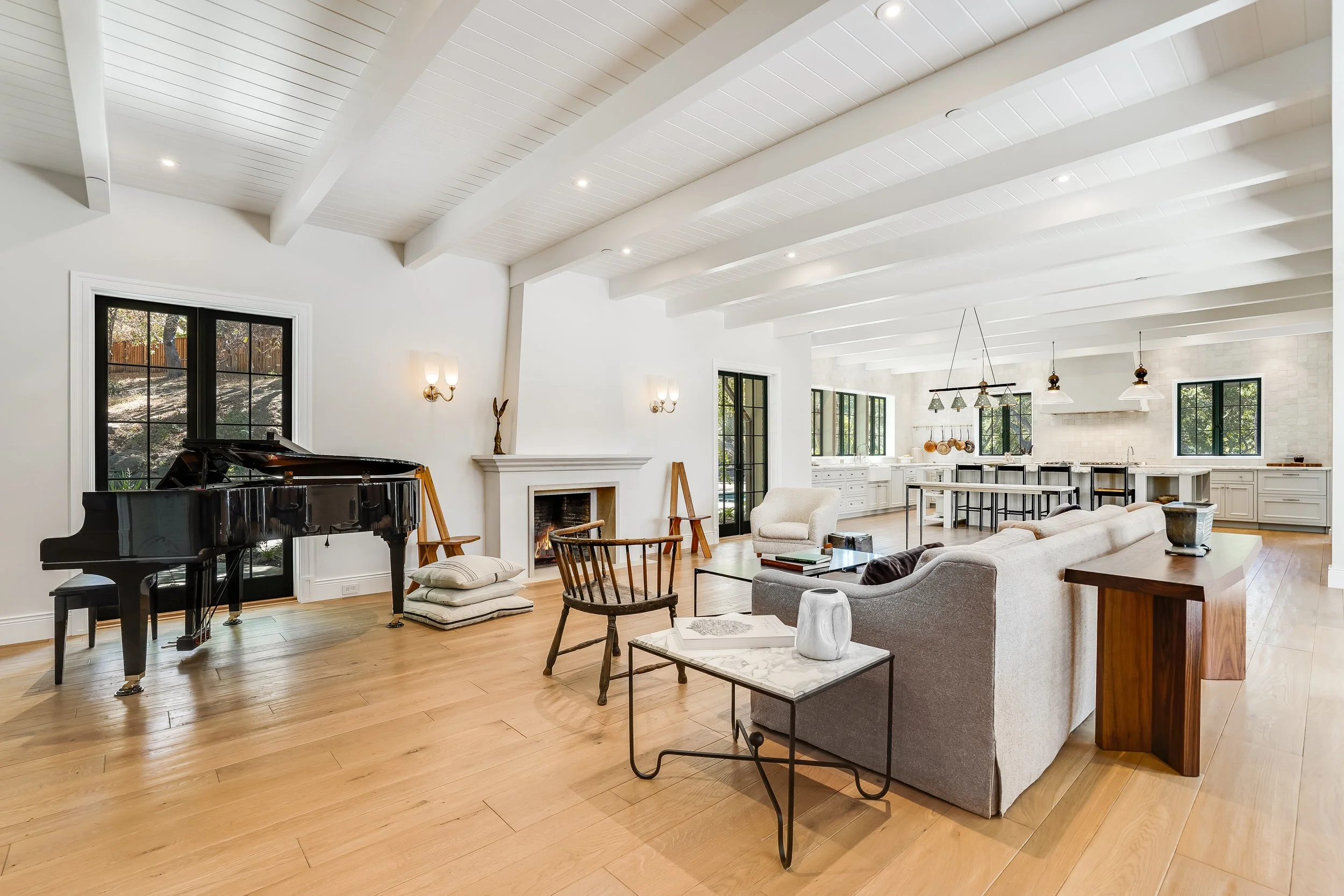 Open-plan living room with a white beamed ceiling, grand piano, fireplace, and a kitchen with a wood-topped island beyond