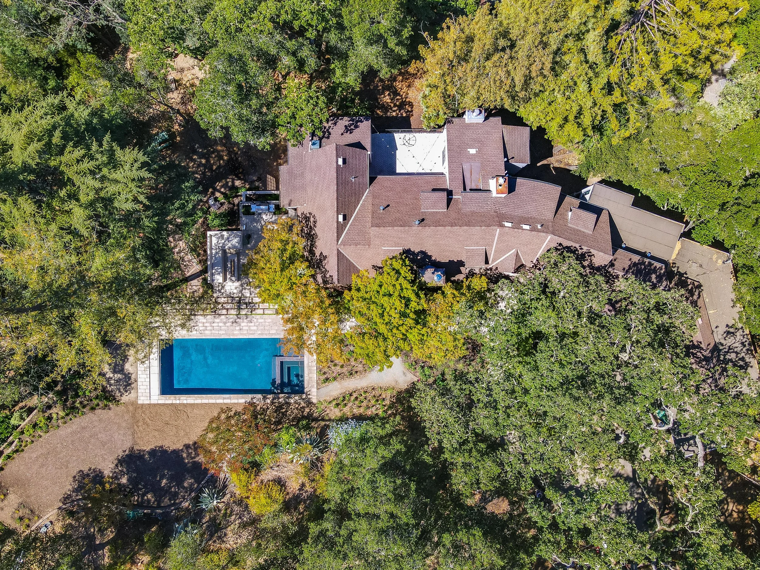 Aerial view of the estate with a dark shingled roof, swimming pool, and surrounding wooded grounds