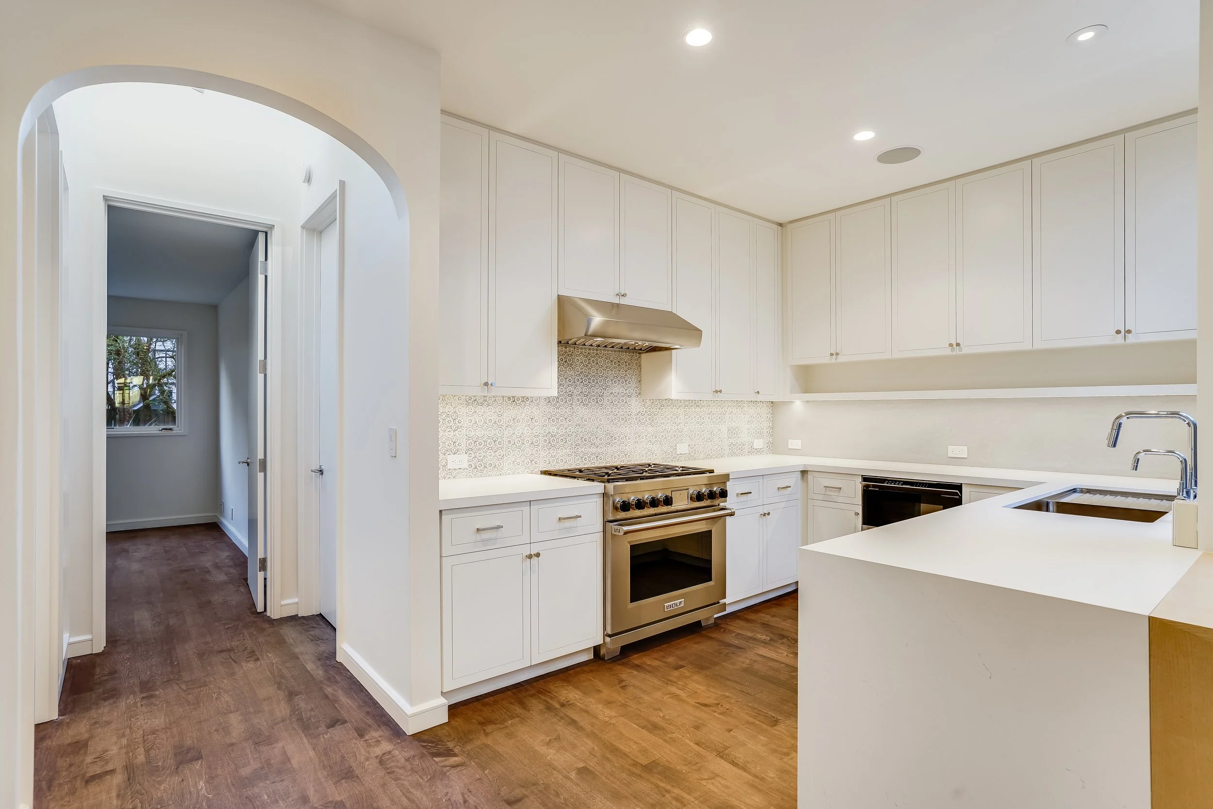 White kitchen with a waterfall-edge island, stainless gas range, hardwood floors, and an archway leading to the hallway