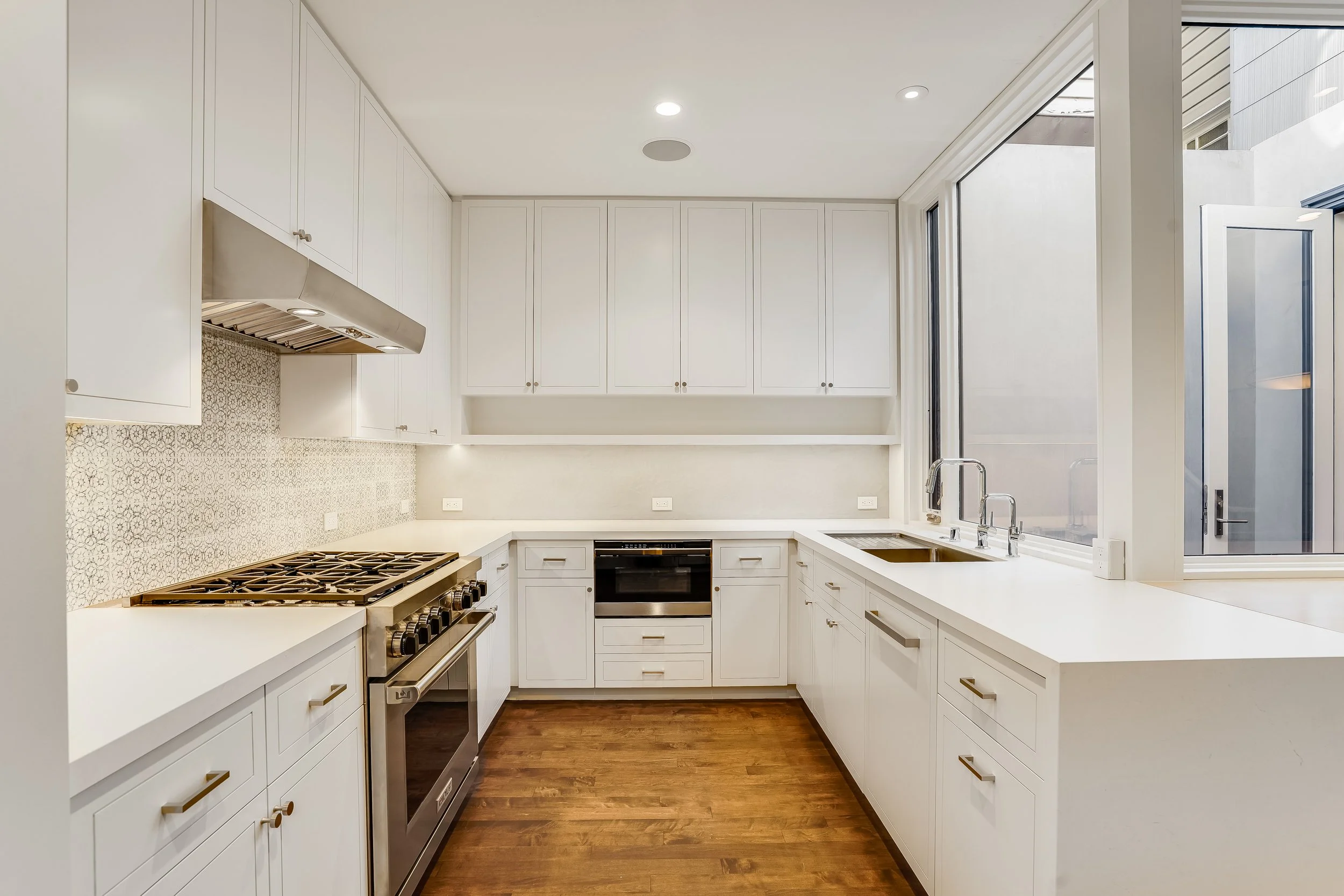 White kitchen with gas cooktop, stainless hood, patterned tile backsplash, and a large window over the counter