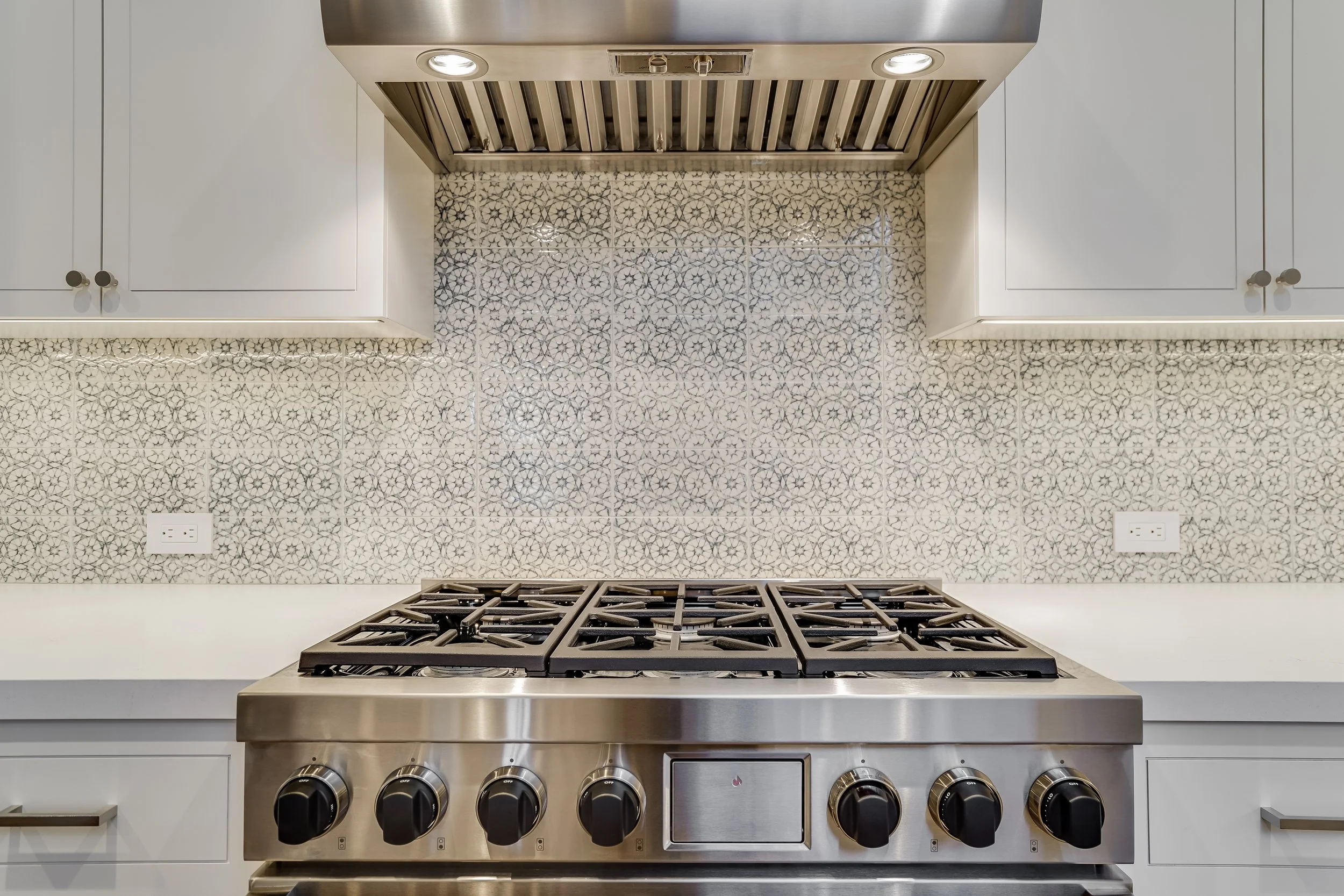 Professional-style stainless gas range with six burners against a patterned tile backsplash and stainless hood
