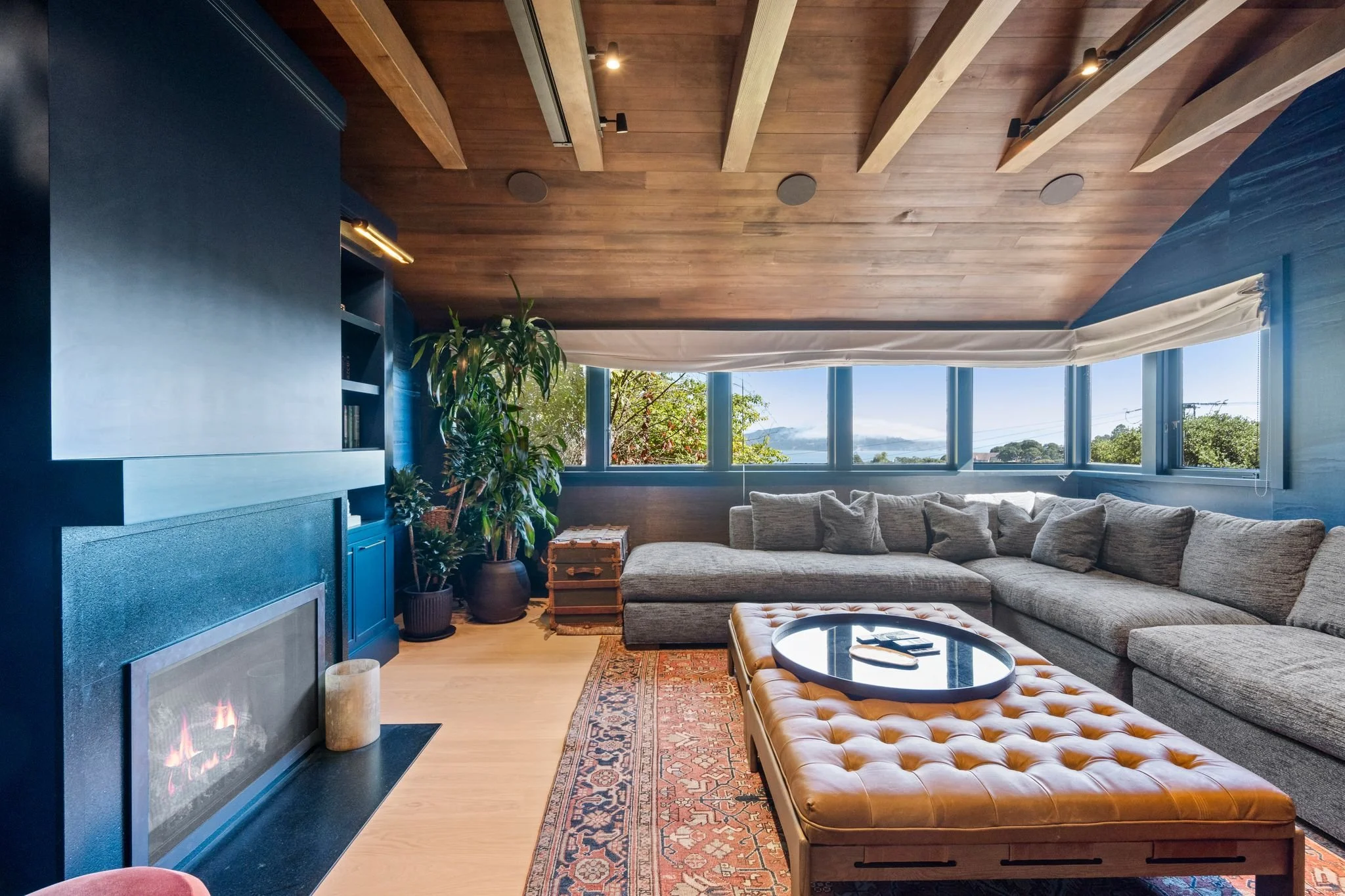Home theater with a linear gas fireplace set in navy walls, gray sectional sofa and tufted leather ottoman, and wraparound windows with bay views