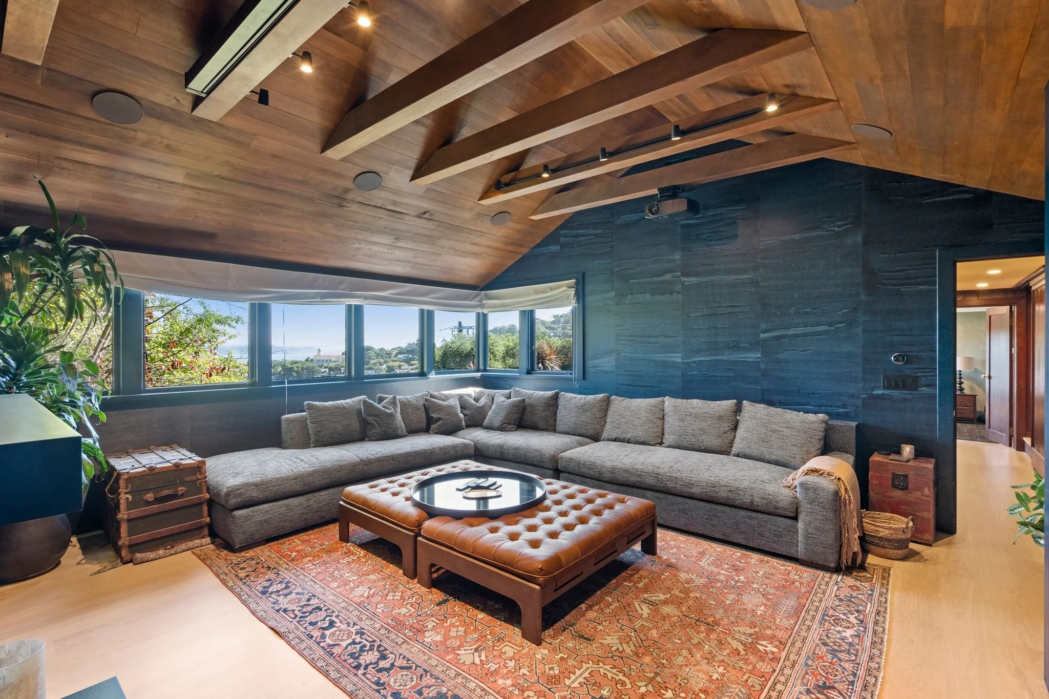 Home theater with a gray sectional sofa, leather ottoman and Persian rug, corner windows with hillside views, and a wood-slat ceiling