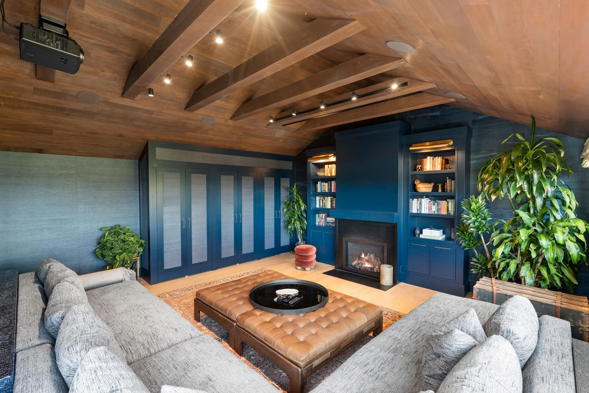 Home theater with navy walls, a corner fireplace and built-in shelving, gray sectional and tufted leather ottoman under a vaulted wood-beam ceiling