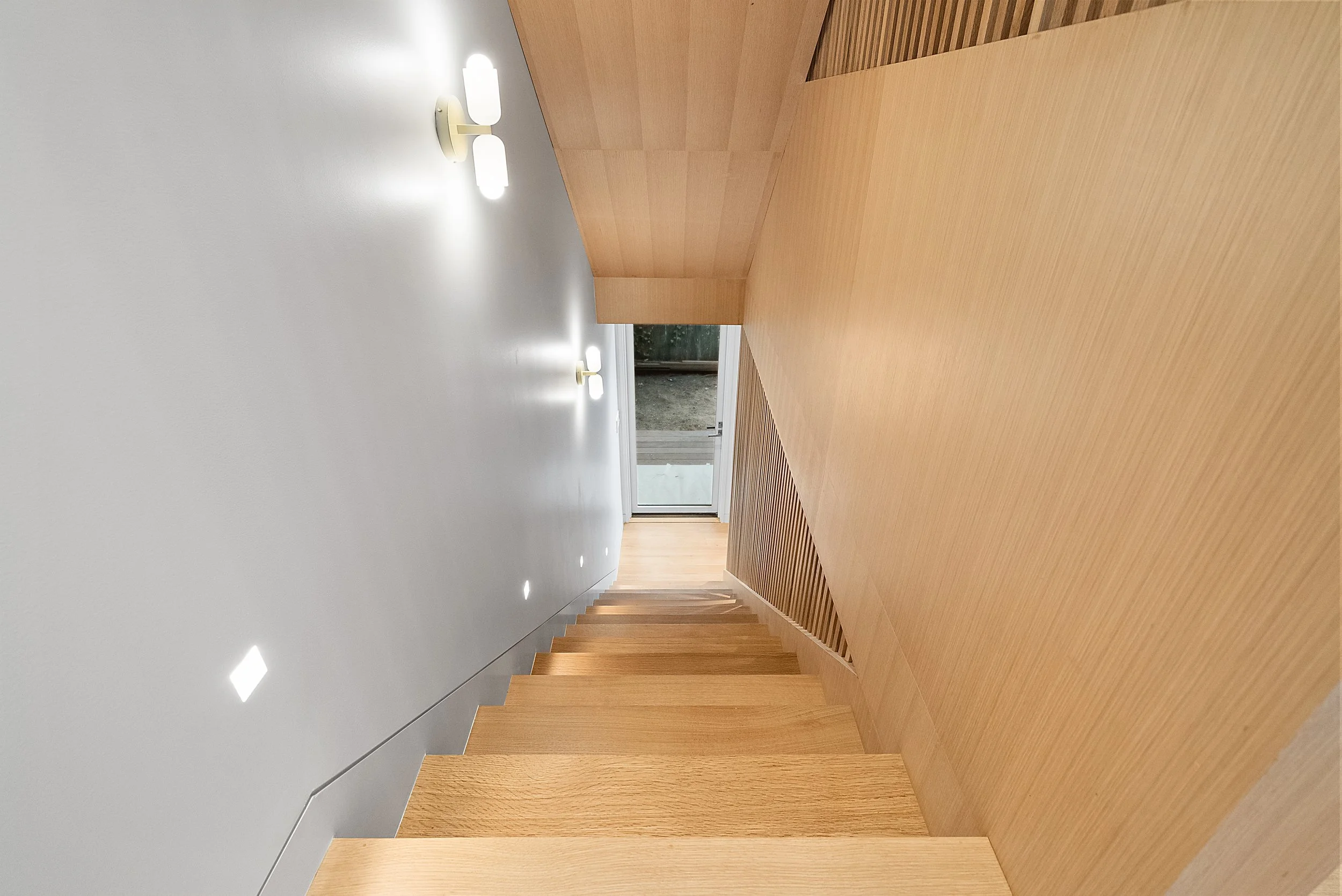View down a light wood staircase with white walls and a wall sconce