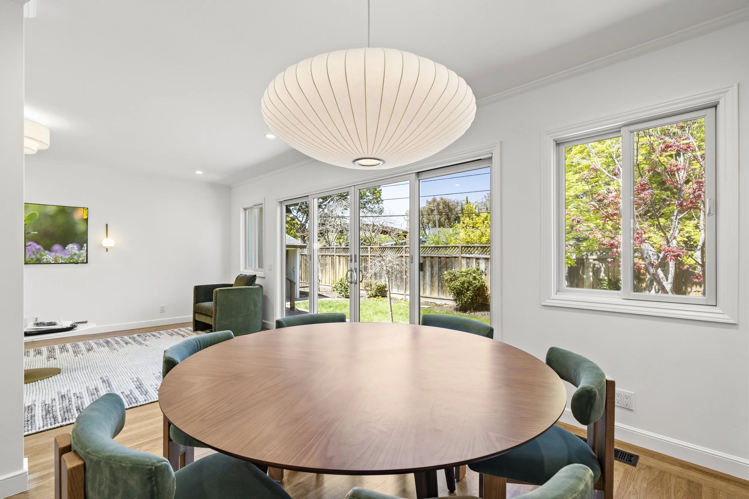Dining area with a round wood table, green chairs, large windows overlooking a garden, and a bubble pendant