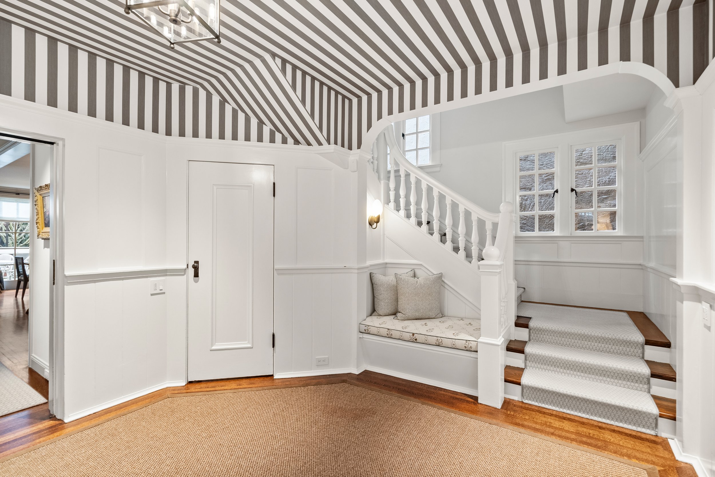 Entry hall with white paneled walls, a striped ceiling, a staircase with a window-seat bench, and a sisal rug