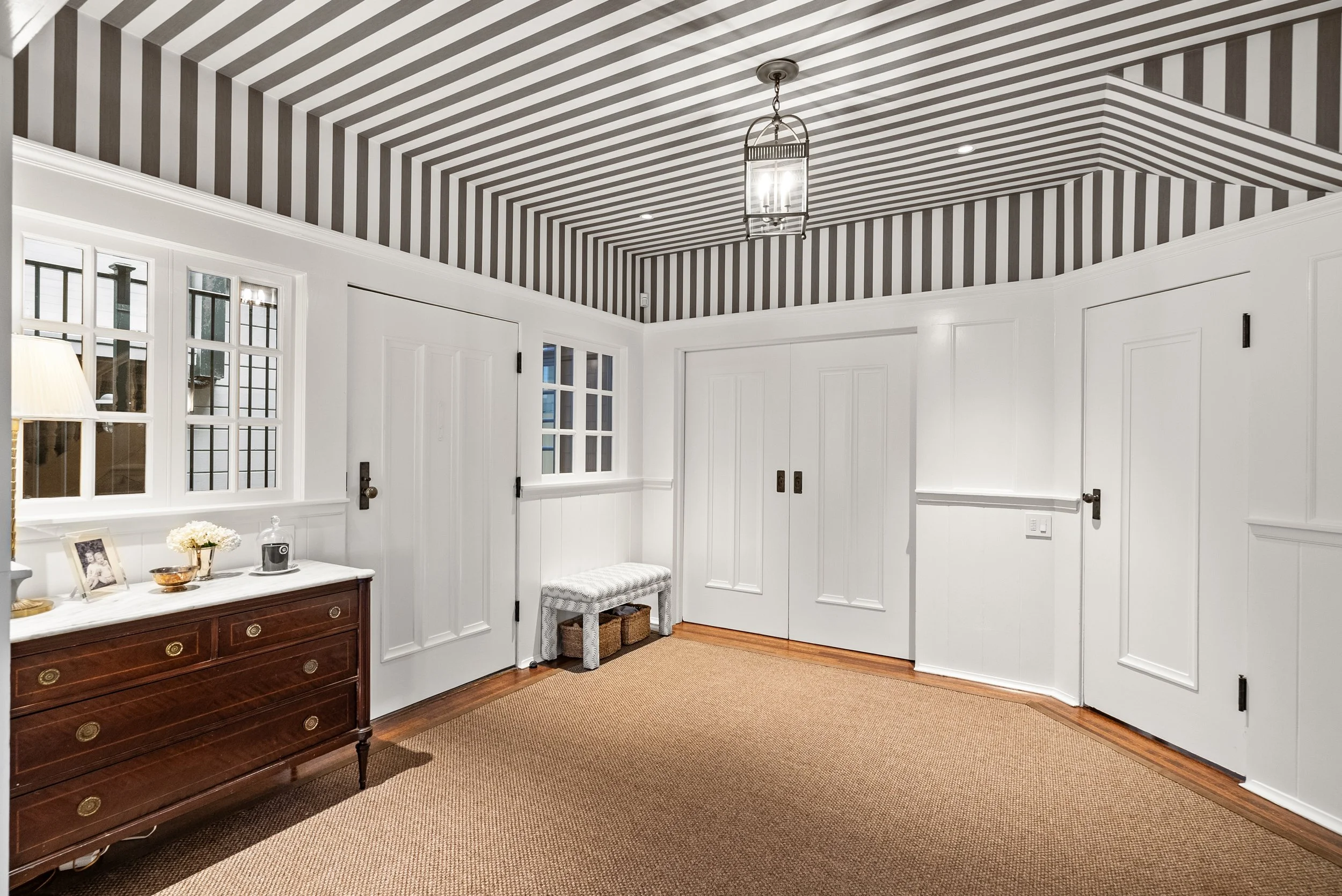 Room with a striped ceiling, white paneled walls, a wood dresser, and neutral sisal carpet