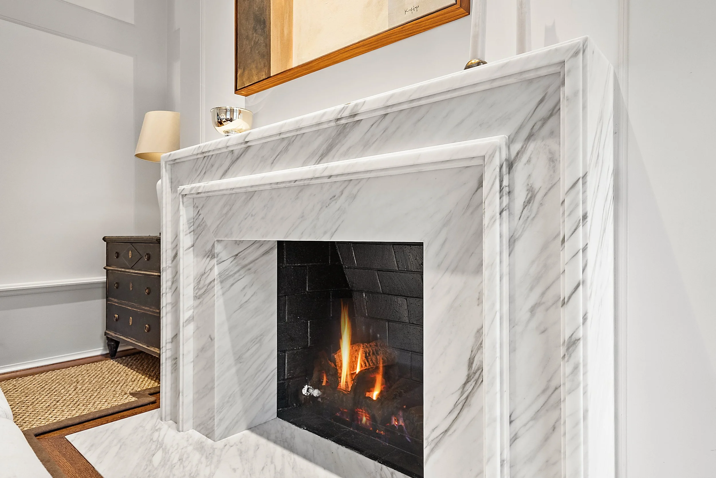 White marble fireplace with a lit fire, a framed mirror above, and a table lamp