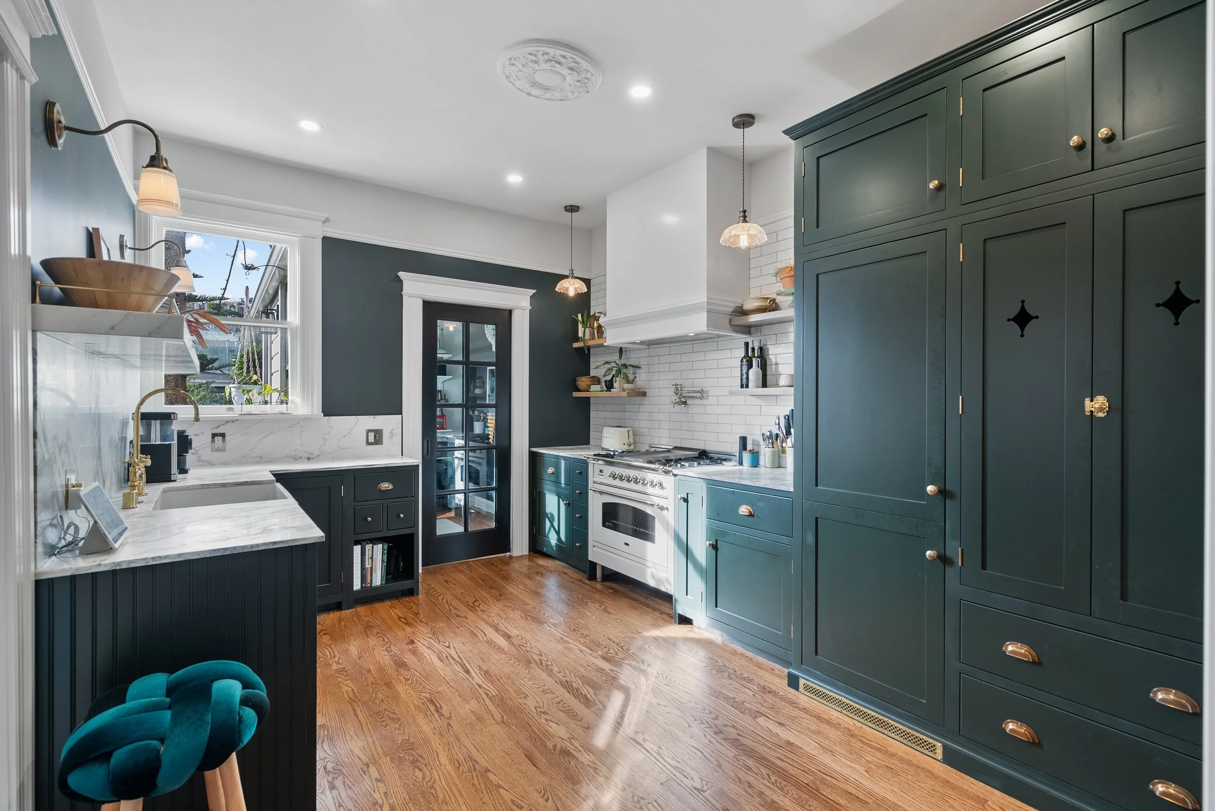 Dark green kitchen with a stainless range, marble countertops, floating shelves, hardwood floors, and a teal bar stool