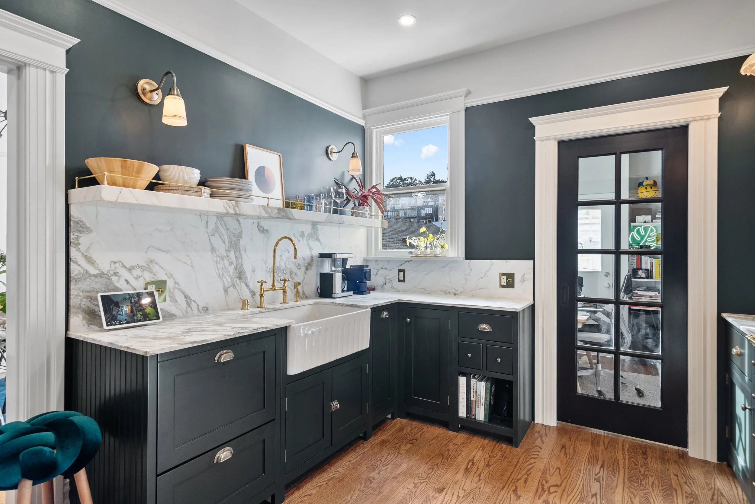 Dark green kitchen with a marble backsplash, a farmhouse sink, a brass faucet, open shelving, and a teal stool