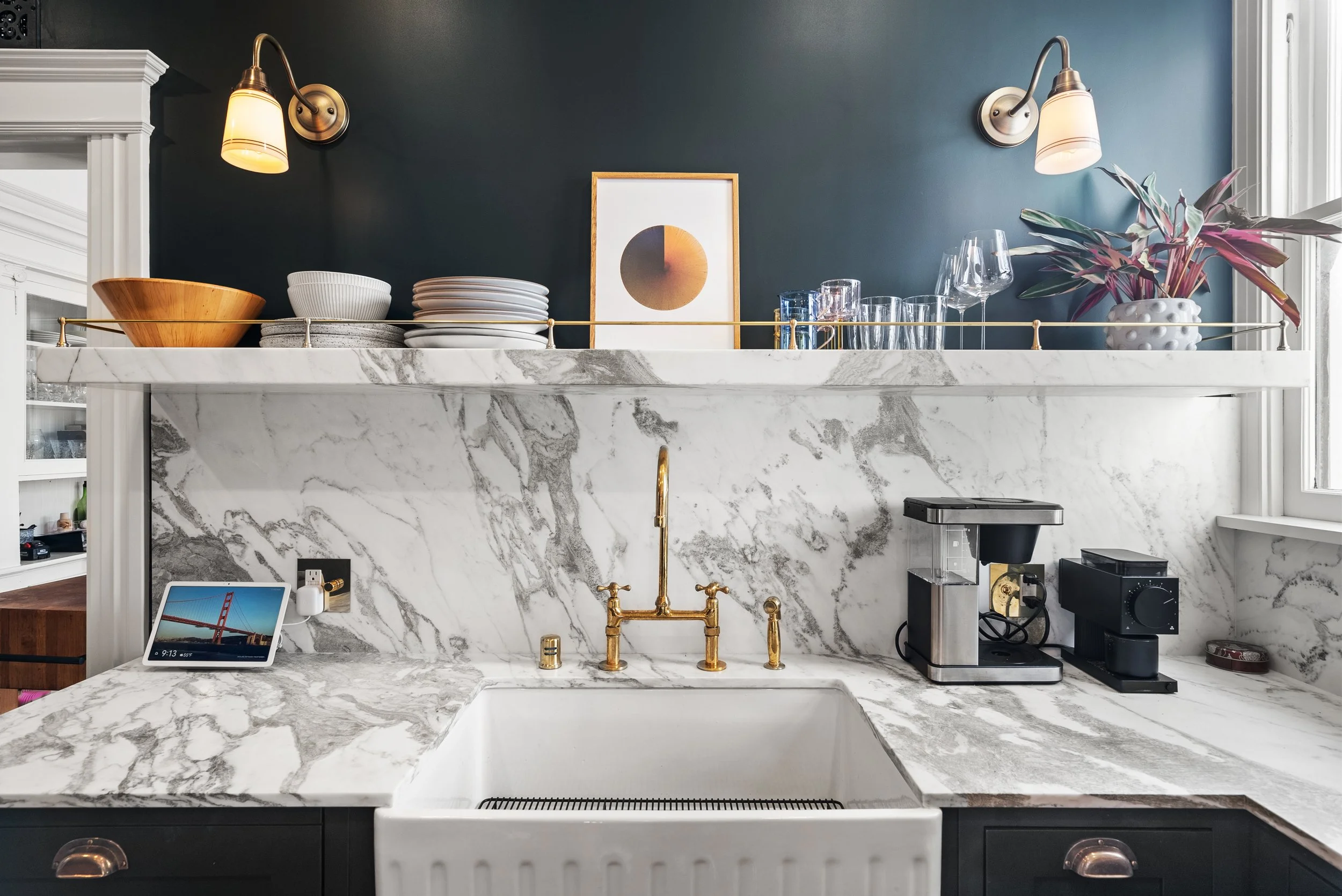 Kitchen with a marble backsplash and countertop, a farmhouse sink, a brass faucet, and open white shelving