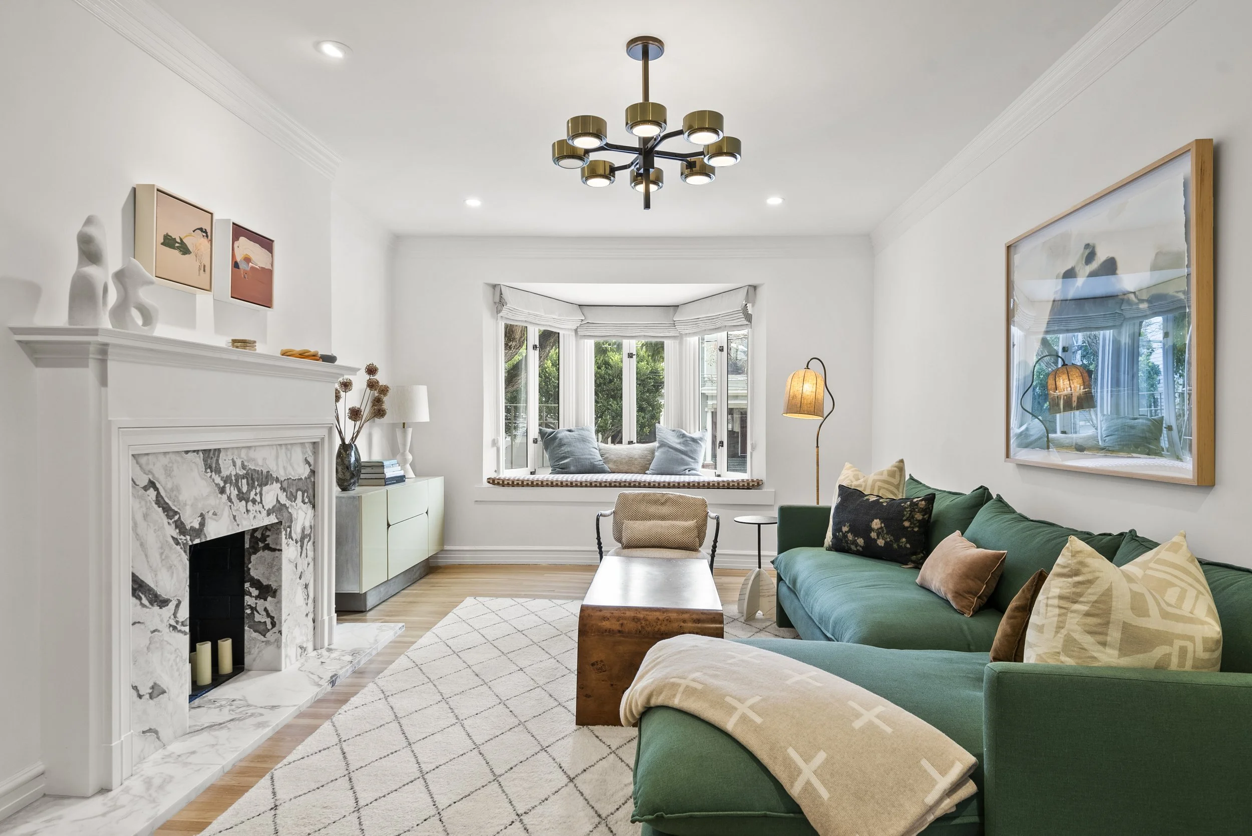 Bright living room with a green sectional sofa, marble fireplace, bay window seat, and framed artwork
