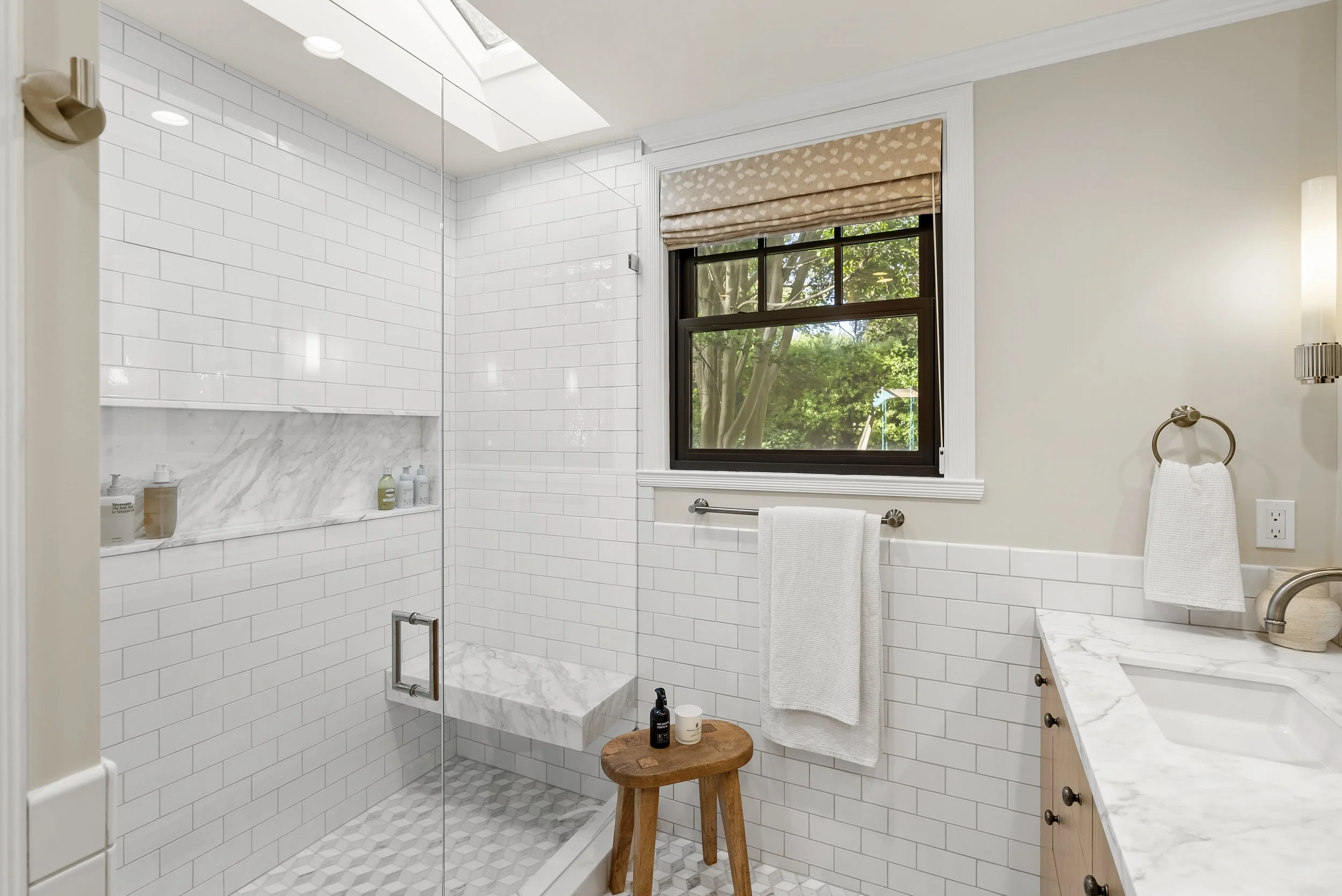 Walk-in shower with white subway tile and a marble bench, beside a soaking tub and a wood stool