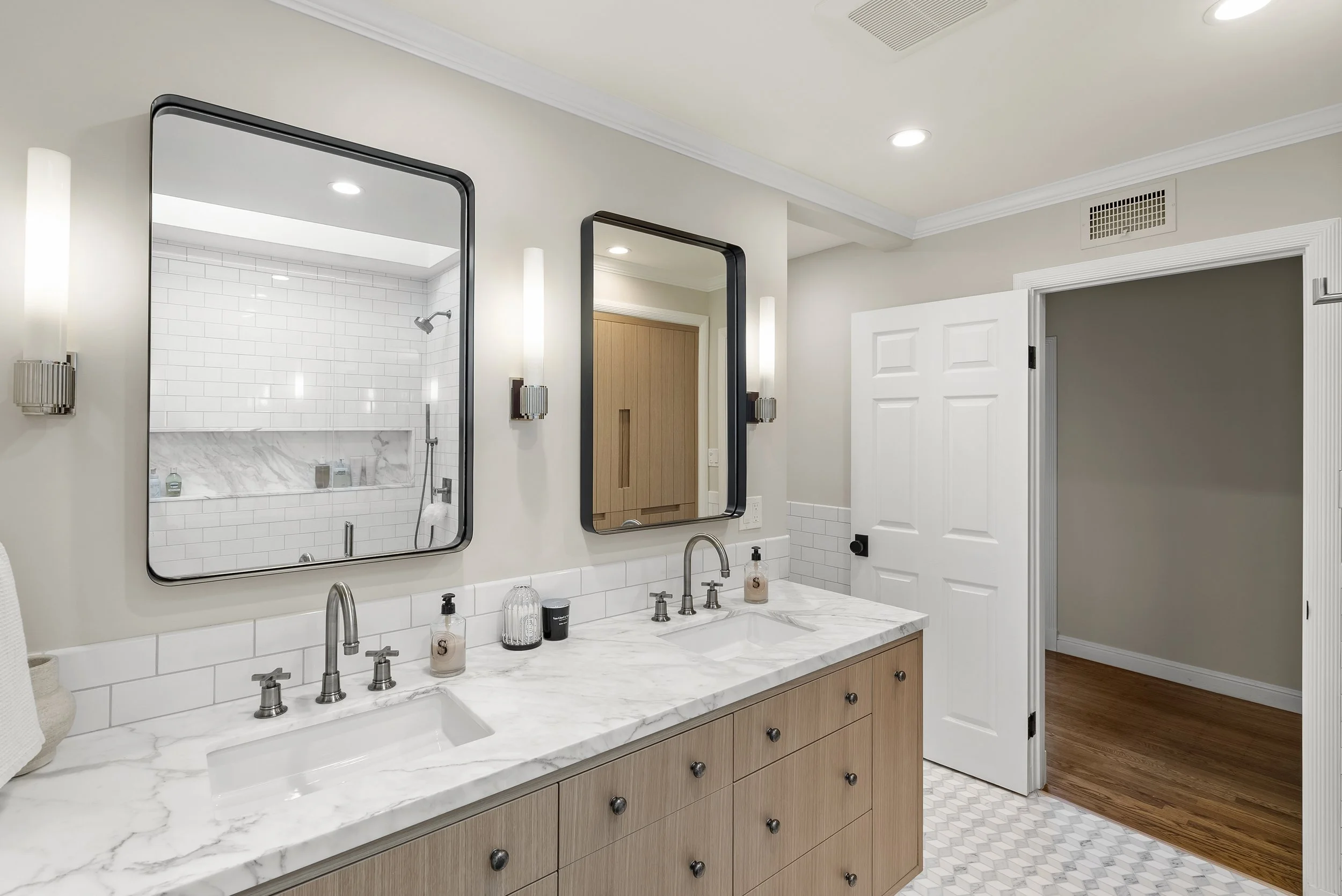 Bathroom with a wood double vanity, marble counter, and black-framed mirrors, with a view to a hardwood hallway