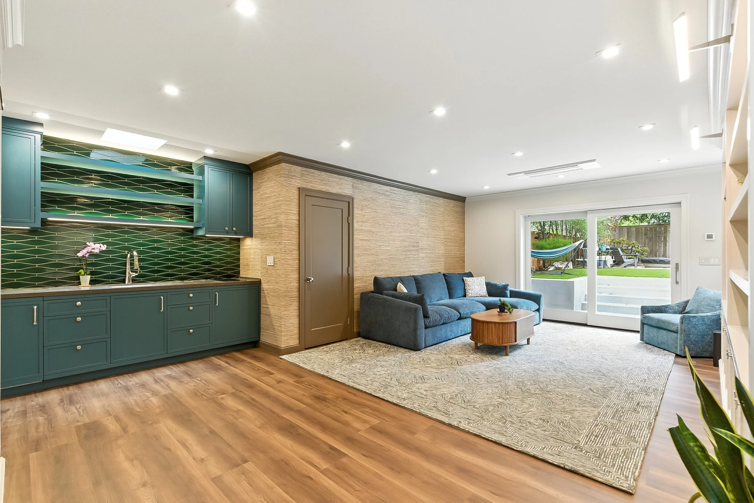 Open living area with a navy wet bar, green tile backsplash, a blue sofa, an area rug, and hardwood floors