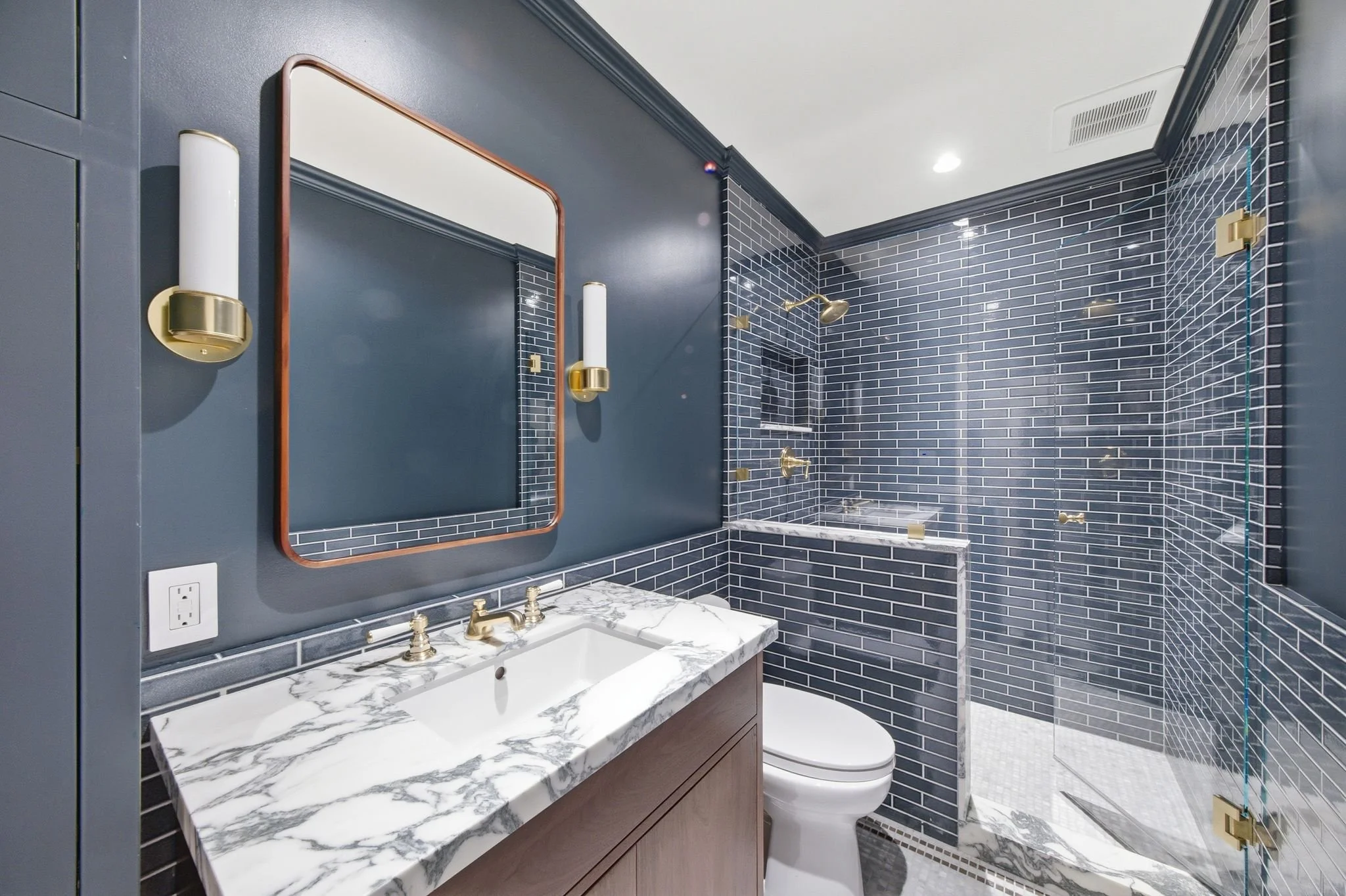 Bathroom with navy walls, a blue glass subway tile shower, marble vanity, gold fixtures, and a rust-framed mirror