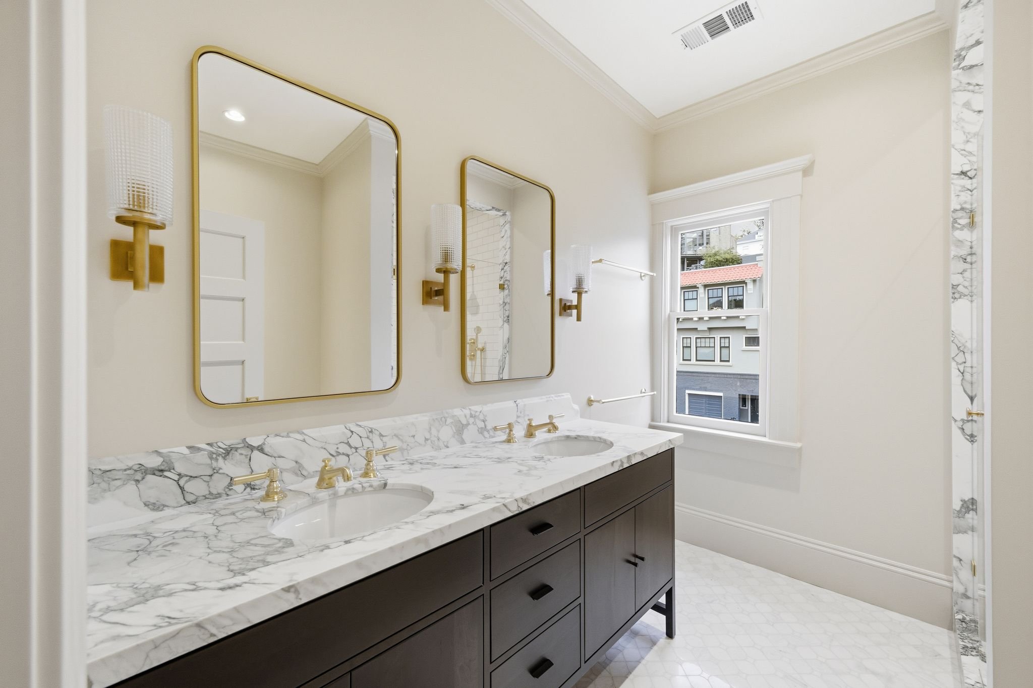Bathroom with a dark double vanity, marble countertop, dual gold-framed mirrors, and brass sconces