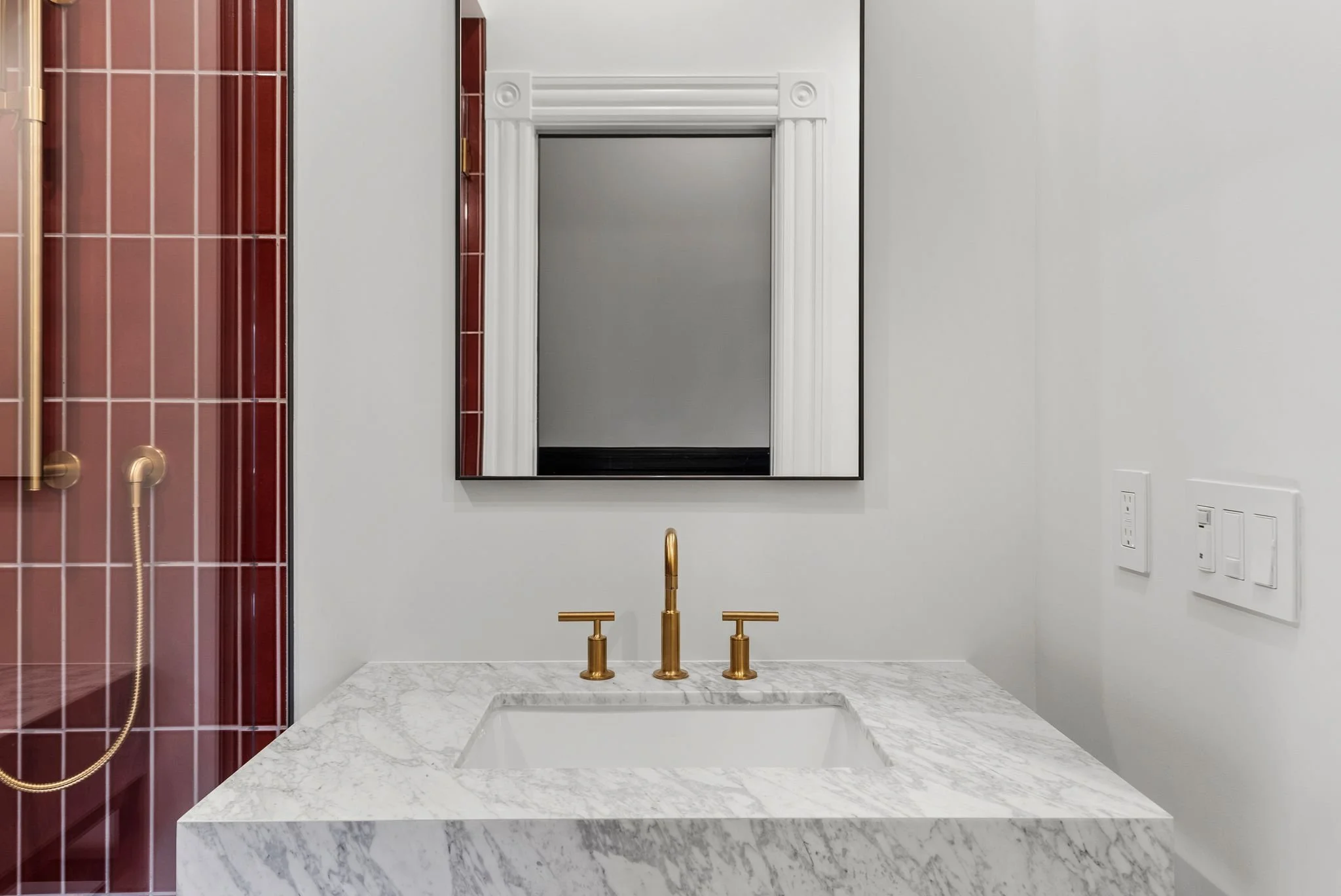Marble vanity top with brass widespread faucet and red tiled shower wall