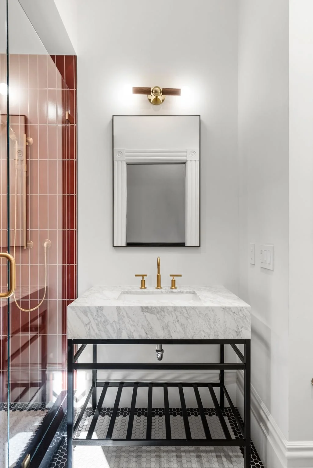 Marble console sink on black metal stand with brass faucet and sconce, red tiled shower behind glass