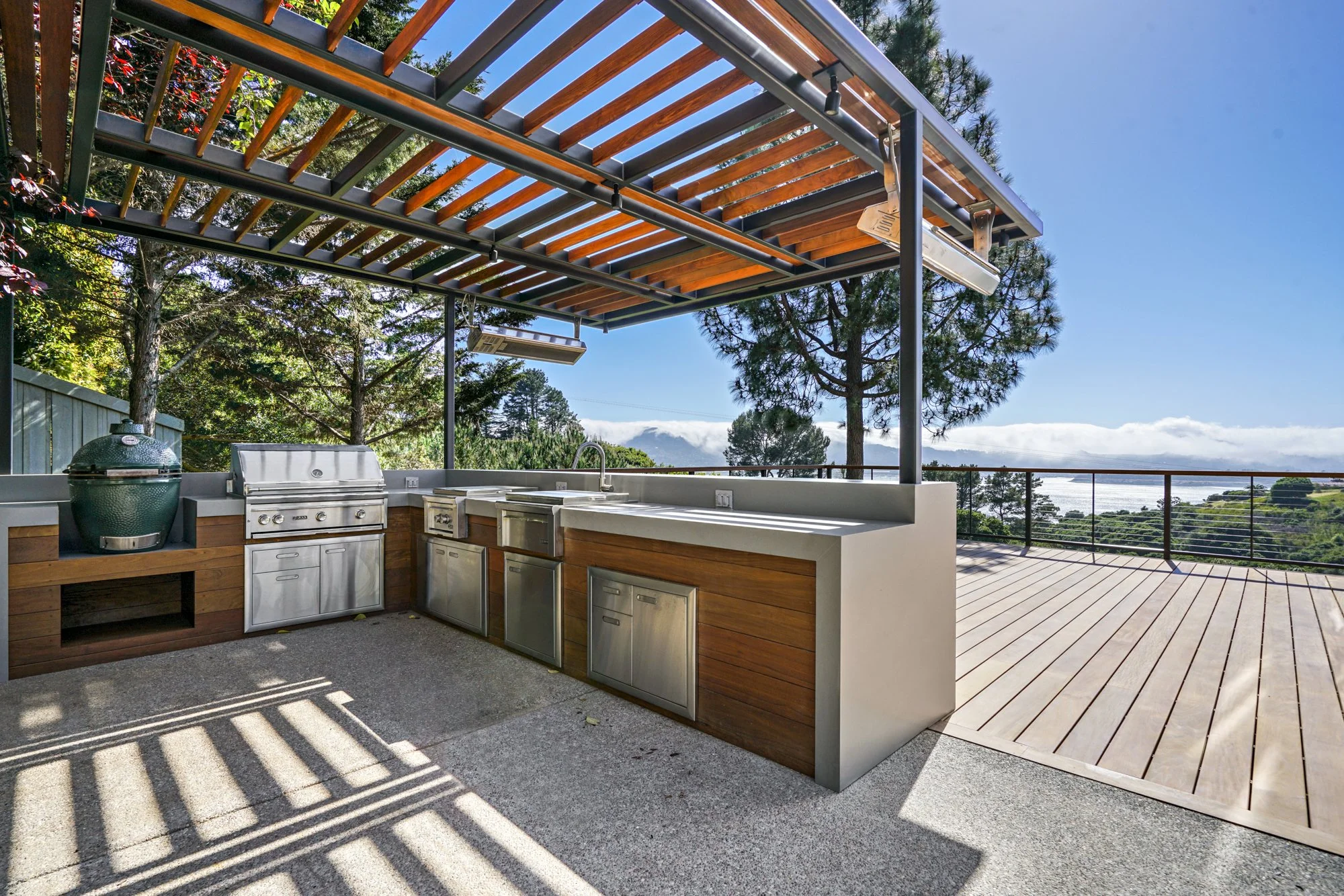 Outdoor kitchen with built-in grill, stainless steel cabinets, and wooden and metal pergola roof. Trees and blue sky in the background, overlooking a scenic landscape.