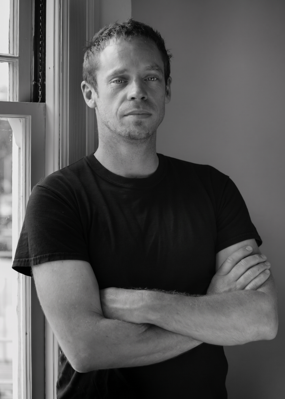 Black and white image of a man with short hair, wearing a black t-shirt, standing with arms crossed near a window, looking directly at the camera with a neutral expression.