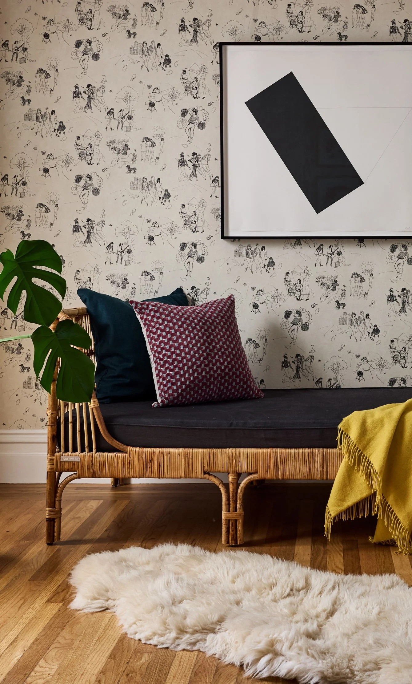 A cozy living room corner with a wicker loveseat decorated with navy and red pillows, a yellow fringed throw, and a fluffy white rug on a hardwood floor. The wall features black and white line drawings of people and activities, with a large white frame displaying abstract art. A large green plant adds a touch of nature to the space.
