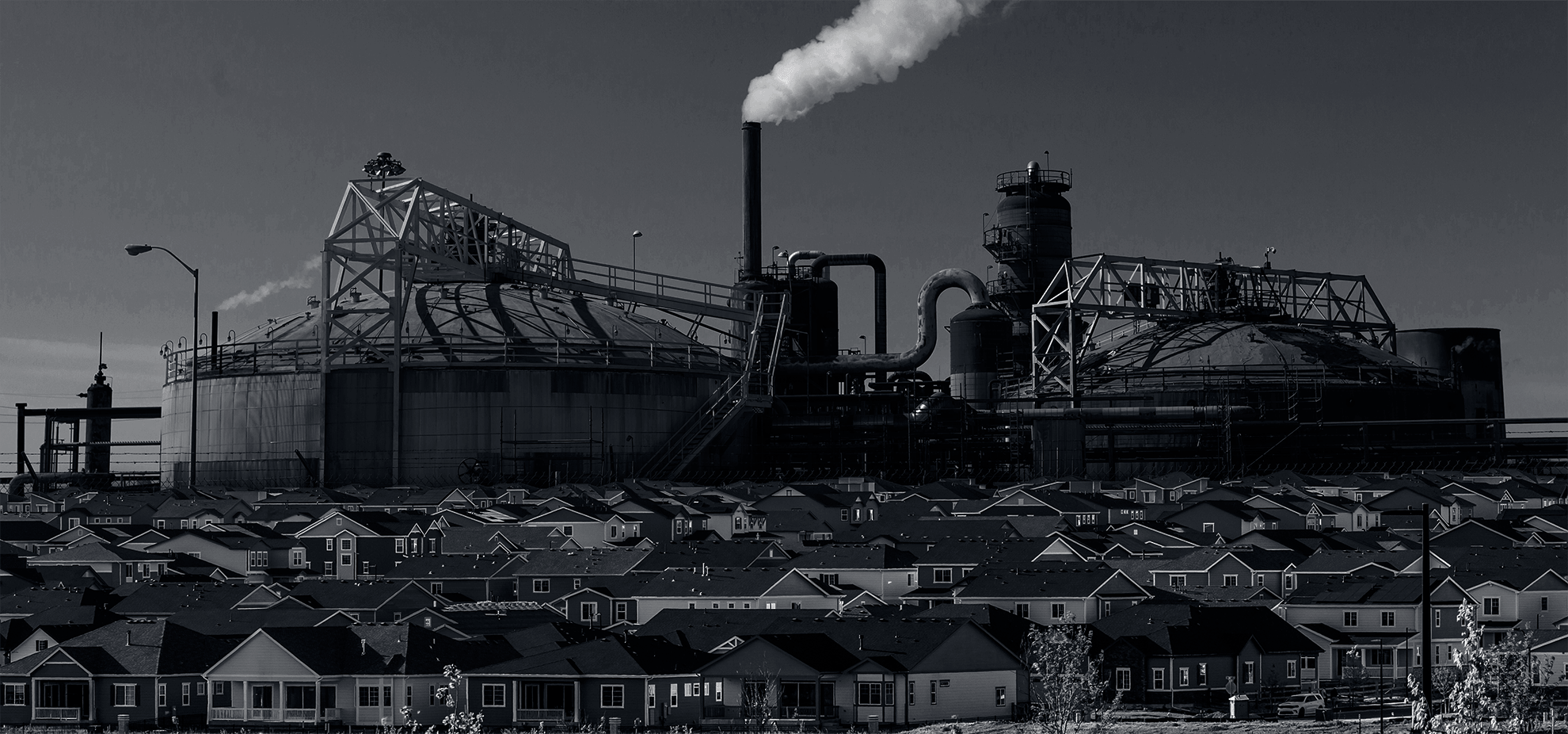 An black and white image of a neighborhood with a large oil & gas refinery superimposed behind it.