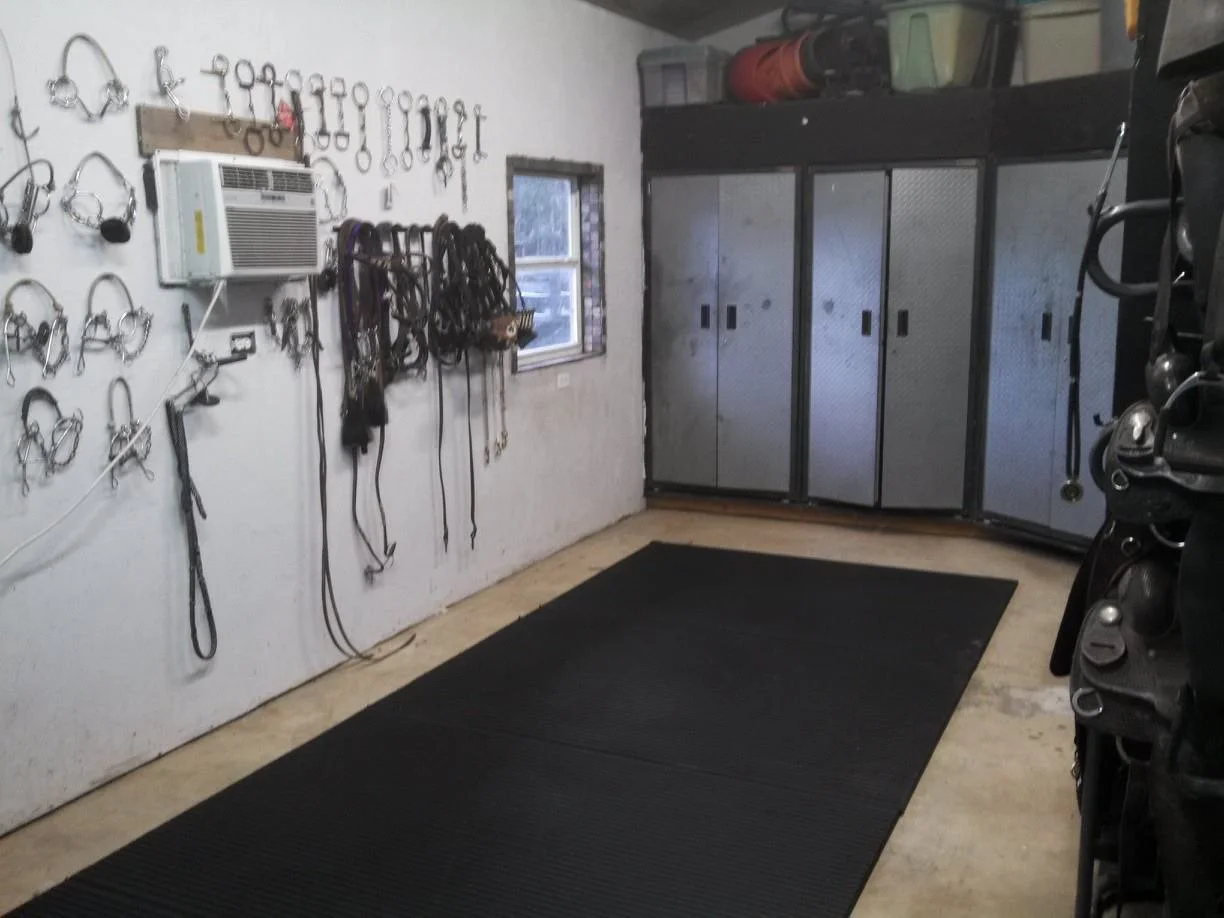 A garage workspace with hooks on the wall holding horse bridles and halters, a window, metal storage cabinets, and several storage bins on top of the cabinets. The floor has a black mat.