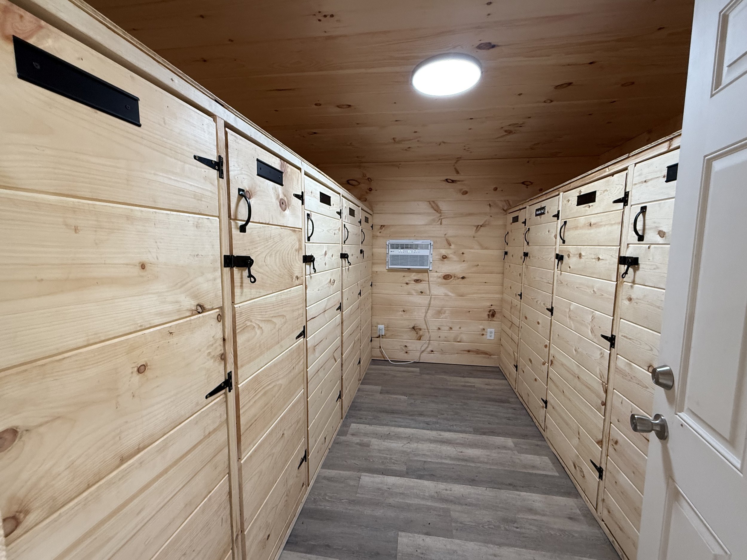 A horse tack room with individual lockers for horse boarding et room with wooden walls and cabinets, an air vent on the back wall, and a wooden floor, illuminated by a ceiling light.