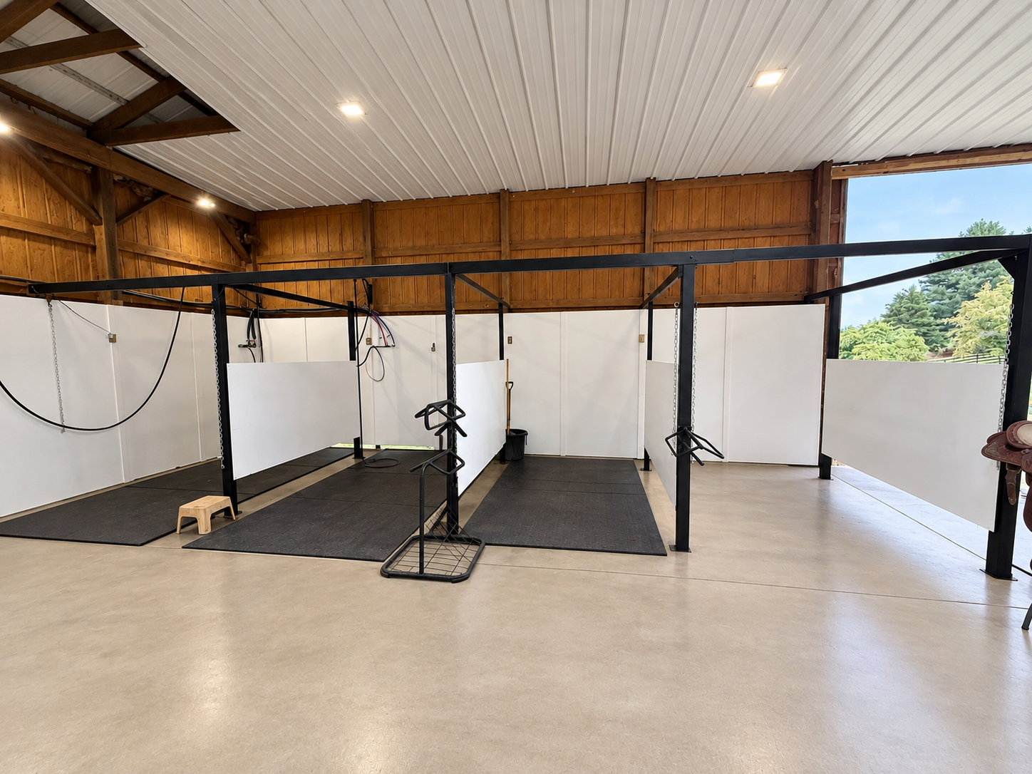 Cross tie wash rack with black metal frame, white wall panels, rubber mats on the floor, and outdoor view through an open section, hot water and cold water for horses and equipment hooks.