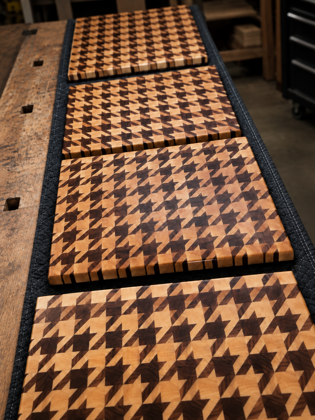 Four wooden cutting boards with a houndstooth pattern in light and dark wood tones, arranged on a black quilted mat on a workbench in a woodworking shop.