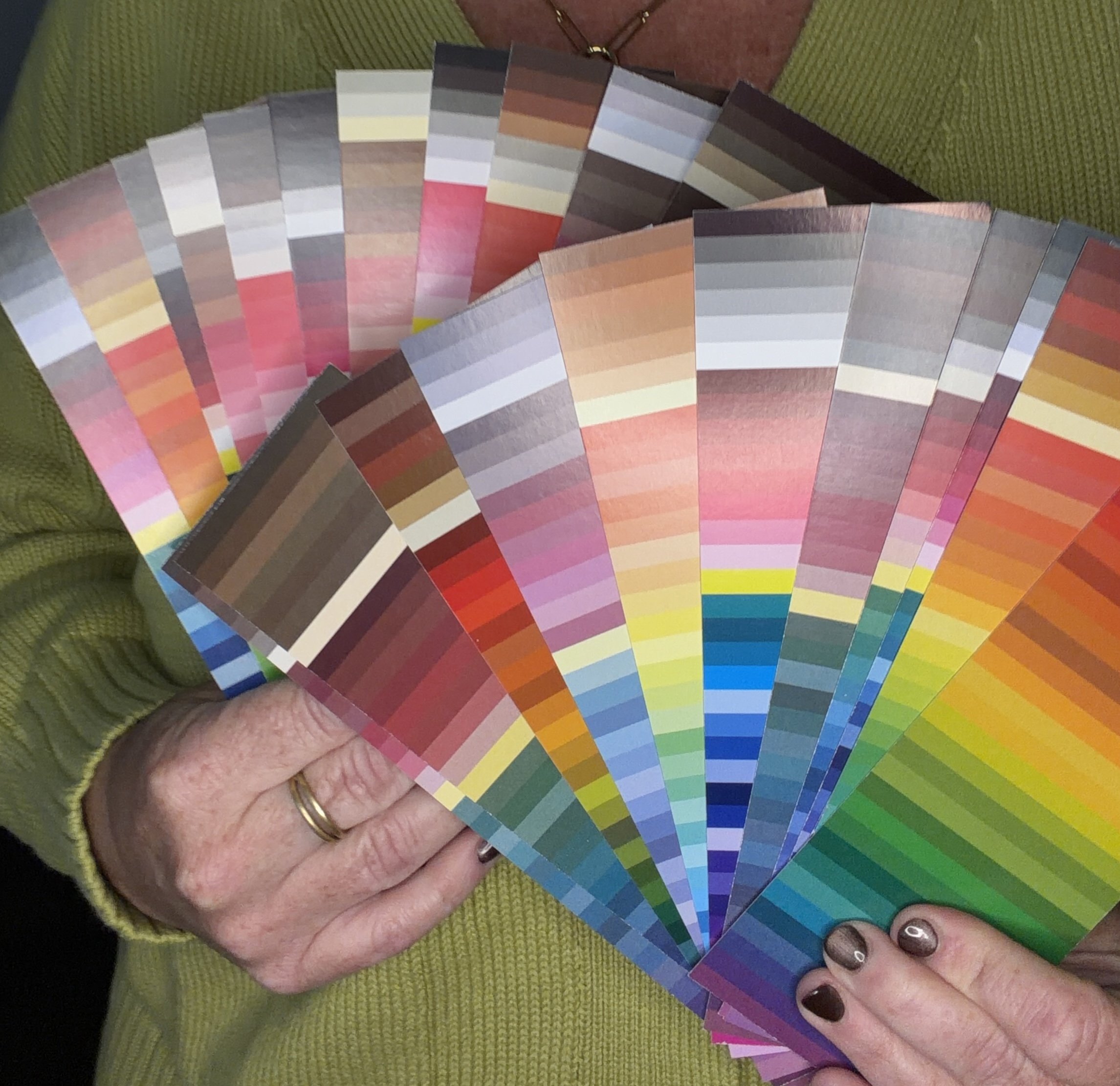 Close-up of a person's hand holding a fan deck of color swatches, with multiple shades arranged in a semicircular pattern, showing a variety of colors from pinks to browns and grays, against a green fabric background.