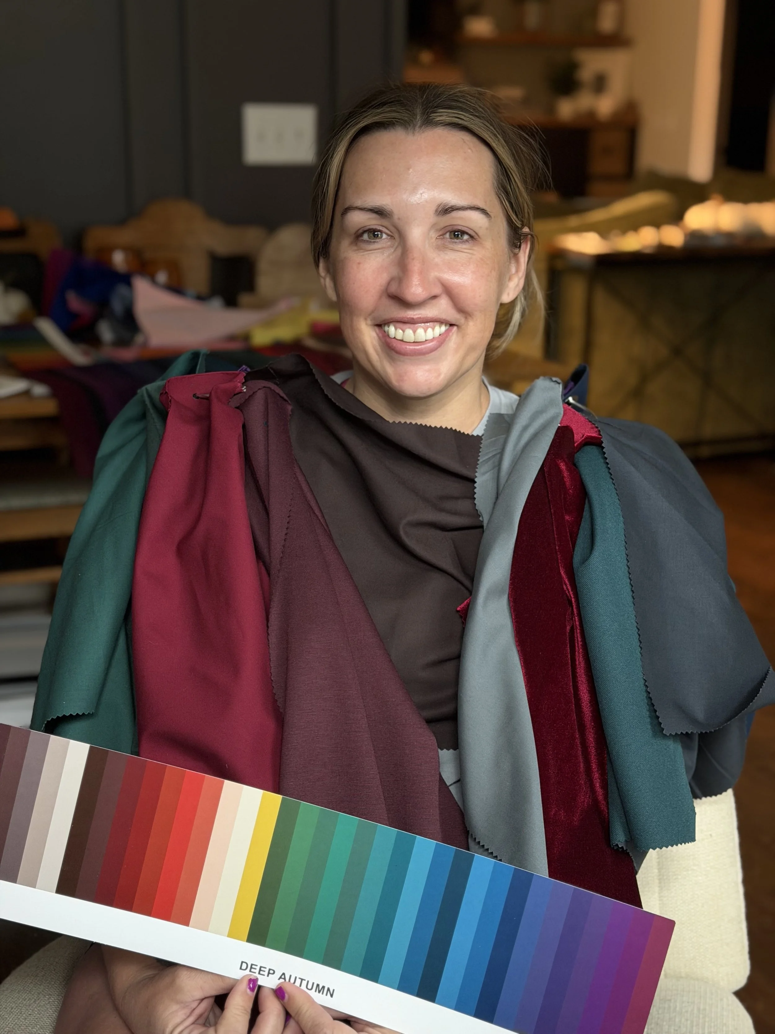 A woman smiling while draped in colorful swatches of fabric in a cozy indoor setting. She holds a color palette labeled "Deep Autumn" in her hand. There are chairs, shelves, and a table in the background.