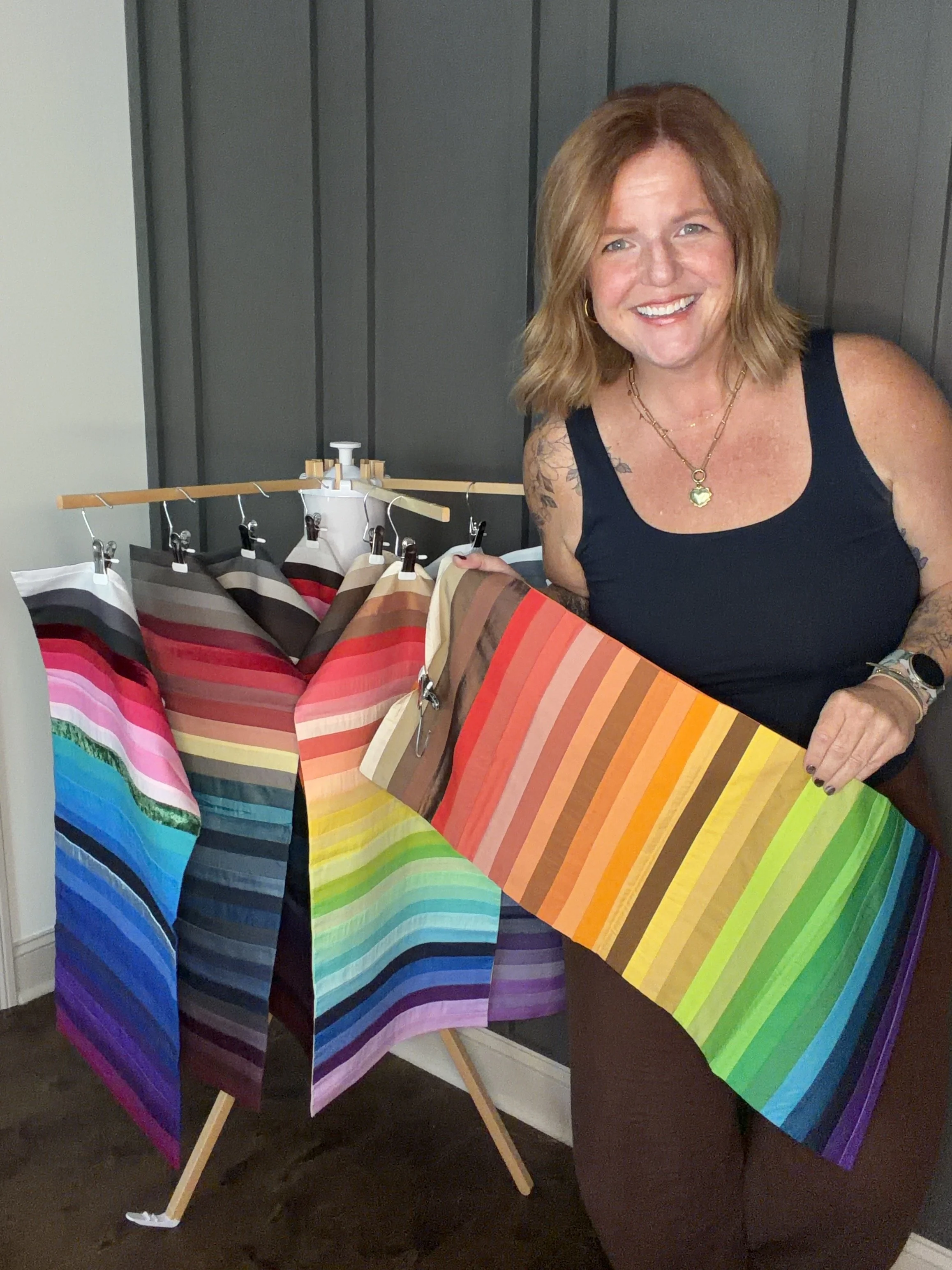 A woman with shoulder-length light brown hair smiling and wearing a black sleeveless top, standing next to a table displaying rainbow-colored fabric swatches on small clipboards and draped over a wooden frame.