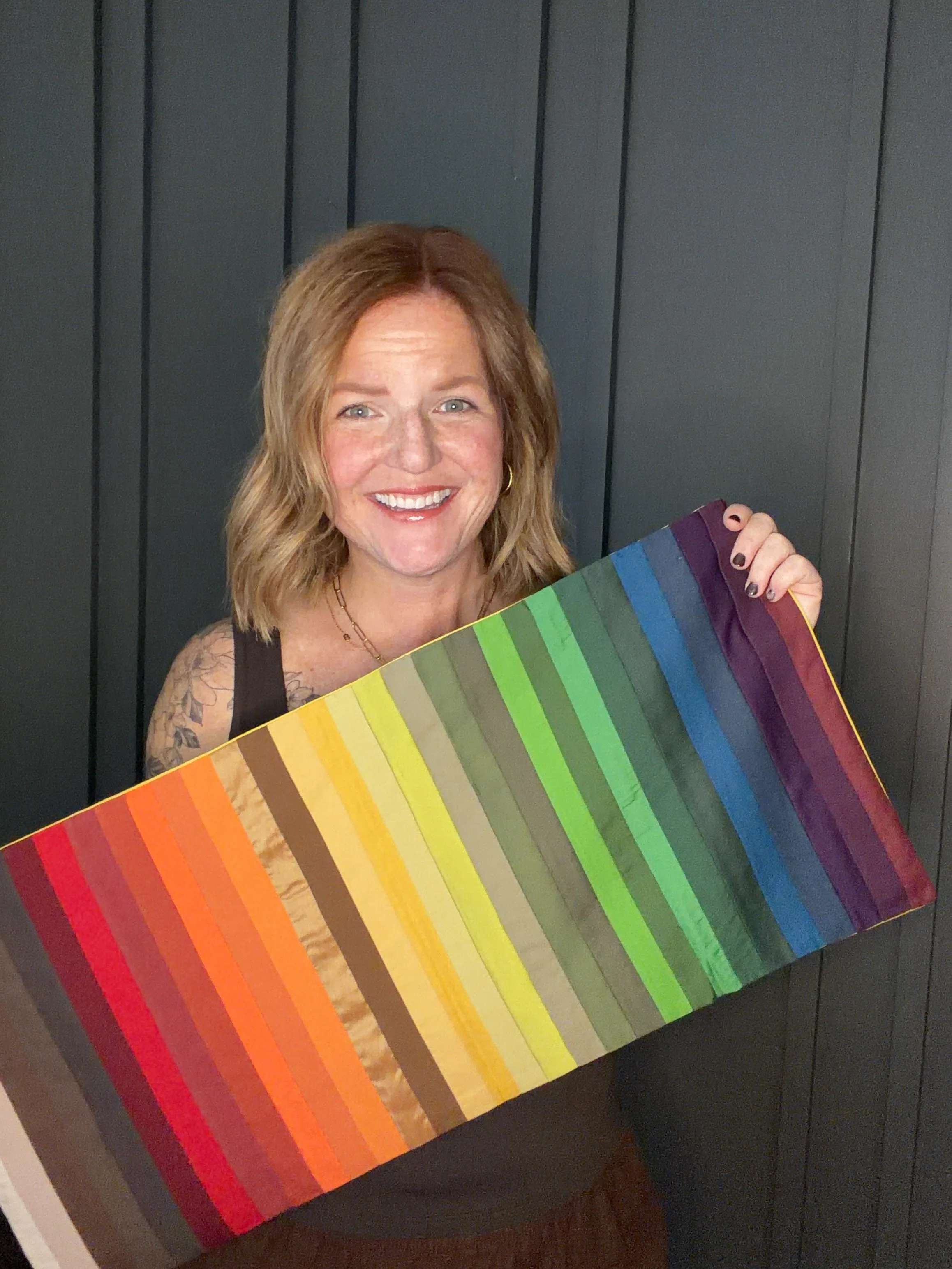 A woman with blonde hair smiling while holding a large rainbow color palette. She is standing in front of gray paneling.