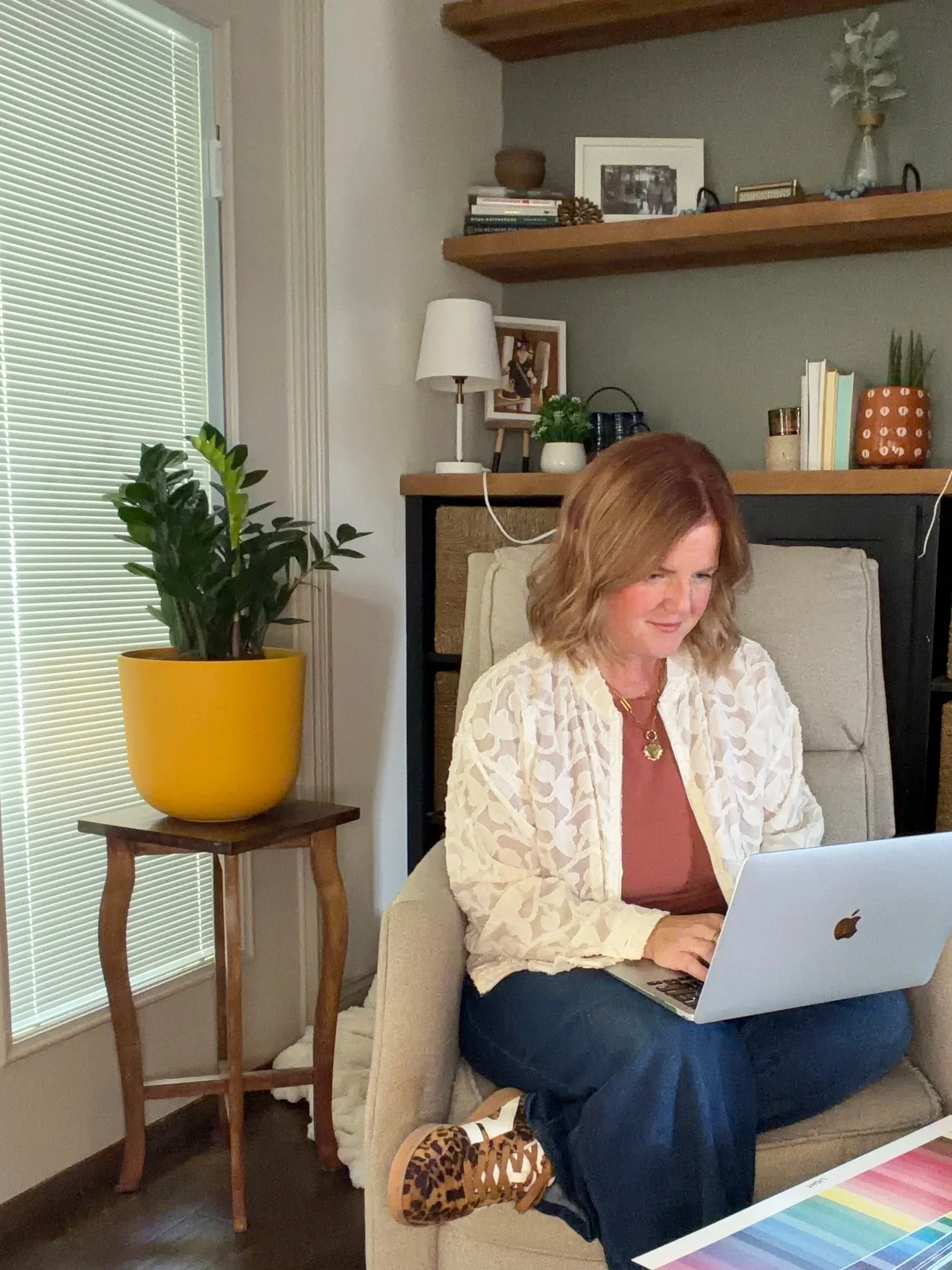 A woman with  hair sitting on a beige armchair, working on a MacBook laptop, wearing a white lace cardigan, a shirt, dark jeans, and leopard print lace-up shoes. Beside her is a wooden side table with a potted green plant in a yellow pot. Behind her, a window with closed blinds, and a wall with two wooden shelves holding books, framed photos, a decorative bowl, and a tall glass vase with white flowers.