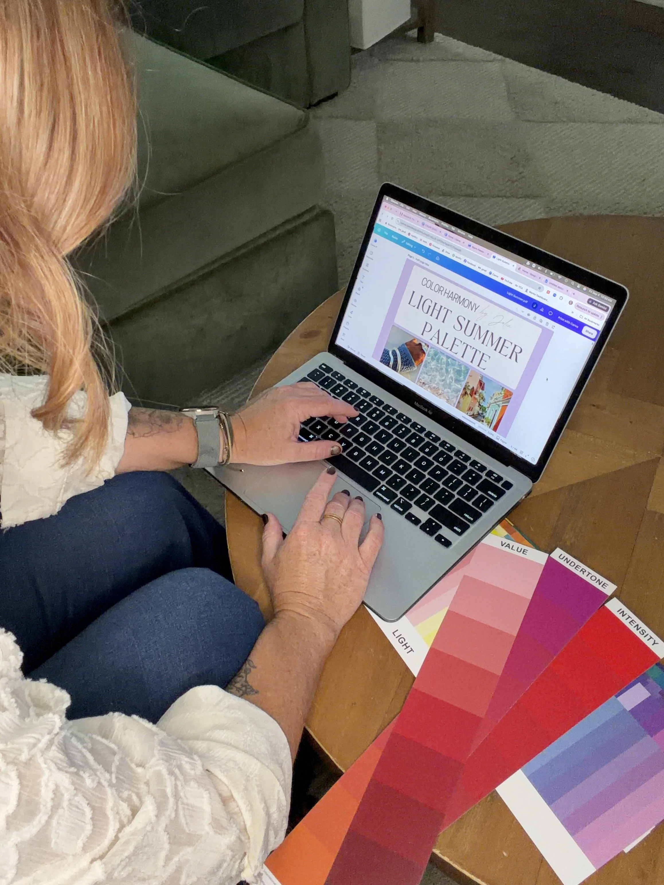 A woman with red hair working on a laptop at a wooden table, surrounded by color swatches labeled with hue aspects like light, value, undertone, and intensity. The laptop screen displays a design project titled "Color Harmony Light Summer Palette."