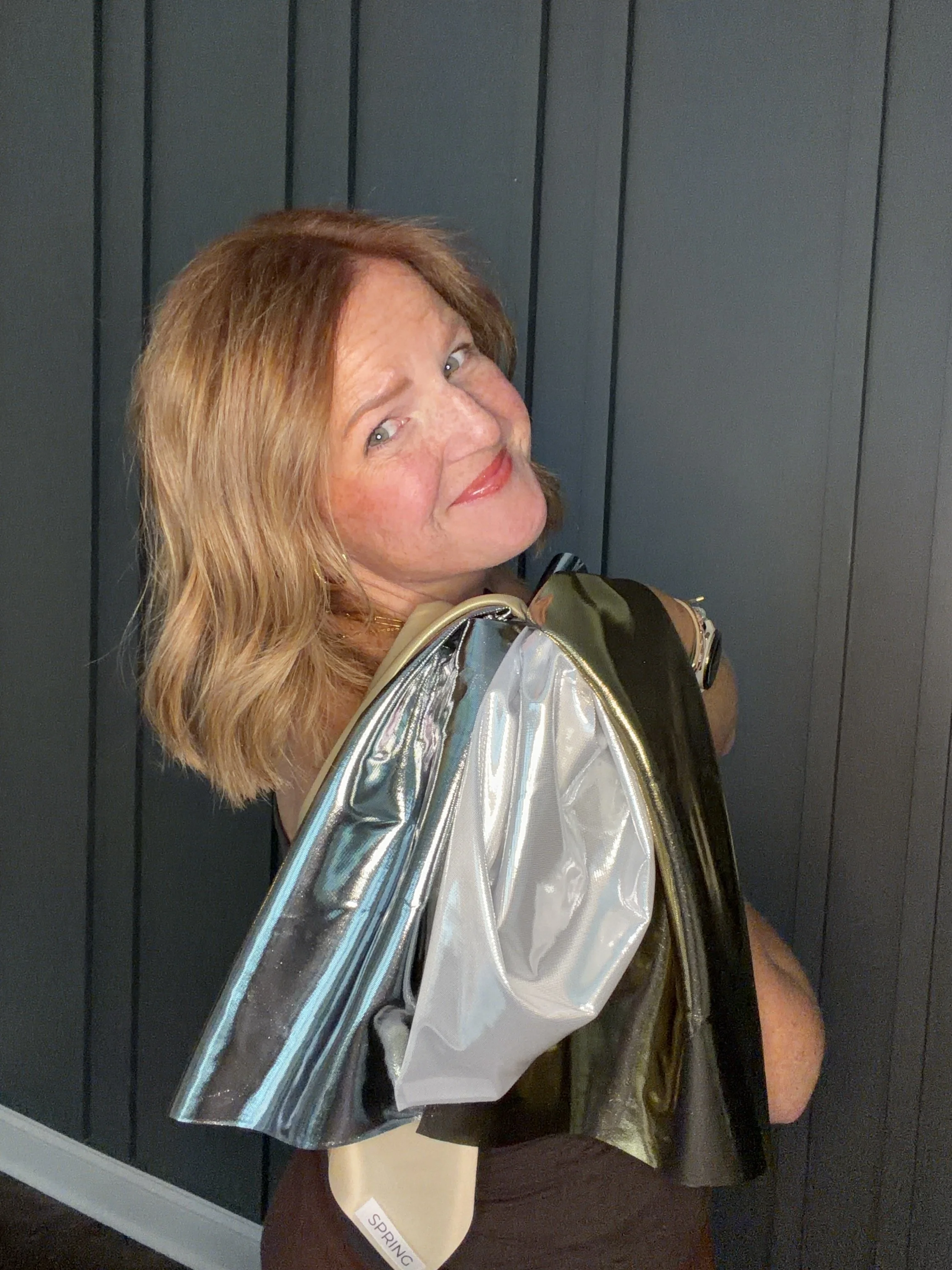 A woman with red hair and green eyes standing in front of a paneled wall, holding  shiny, metallic swatches of fabric, and smiling at the camera.
