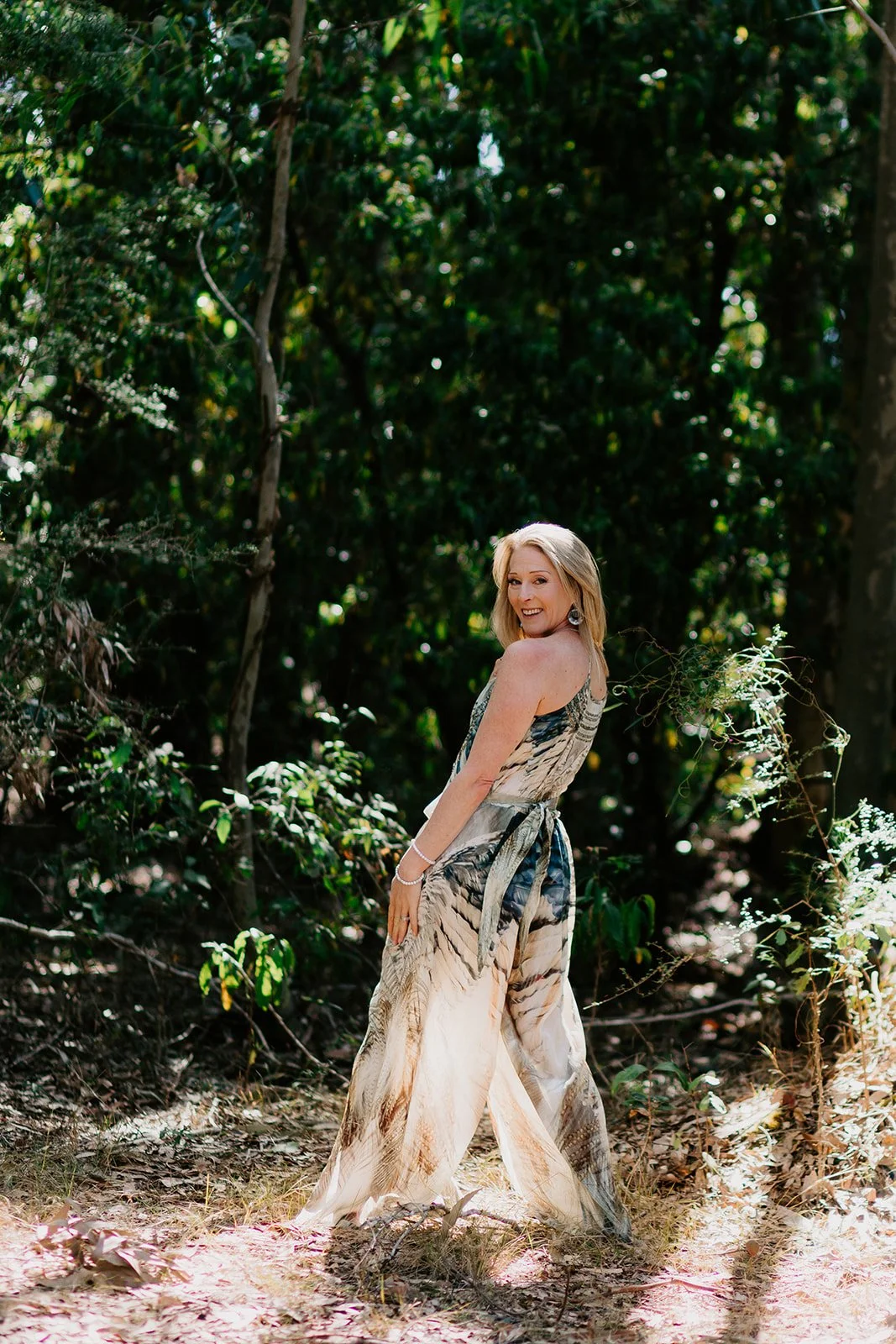 A woman in a flowing dress with a tiger print pattern standing in a wooded area, smiling and looking over her shoulder.