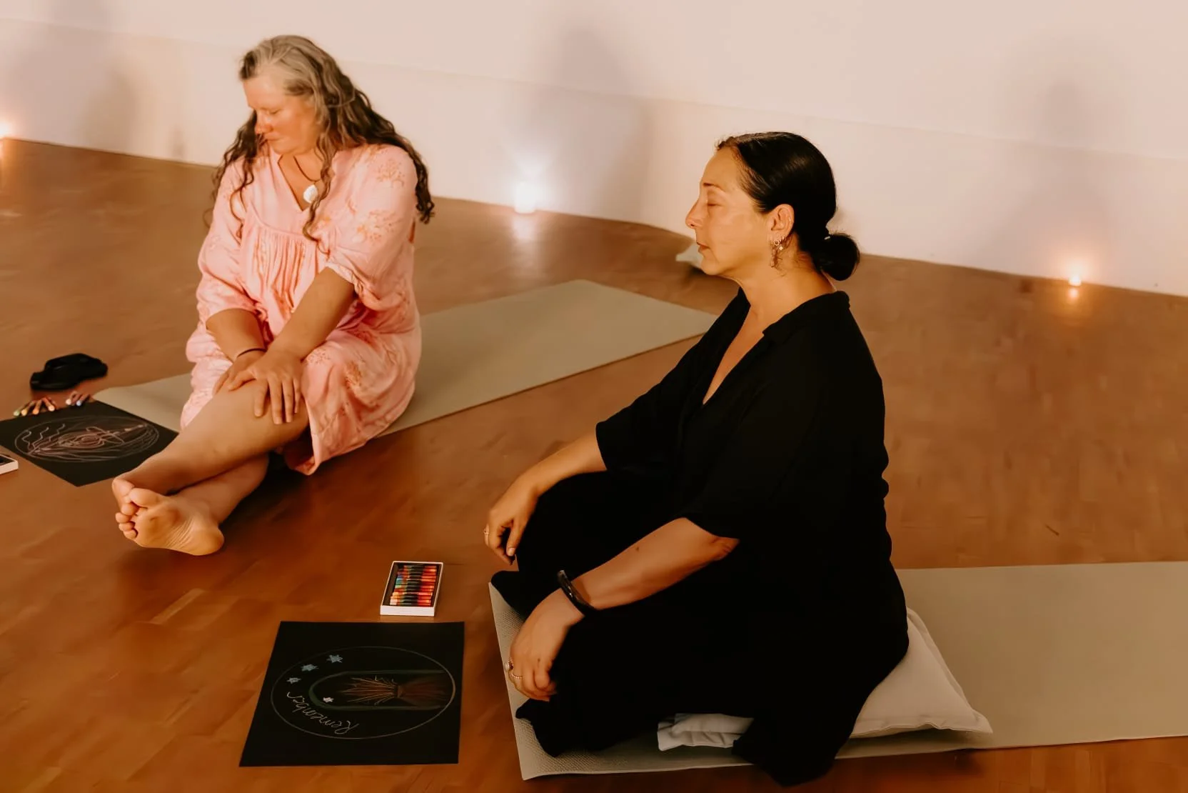 Two women practicing yoga in a softly lit studio with candles on the wall. One woman in a pink dress is sitting on a yoga mat, gently touching her leg. The other woman in a black shirt is sitting on a cushion nearby, watching.