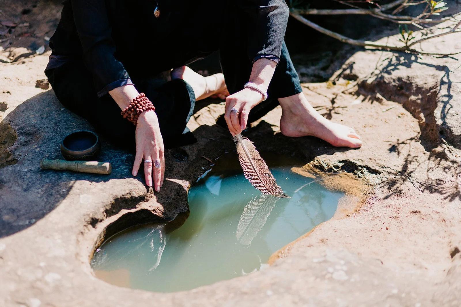 Person sitting on rocks by water, holding a feather. Nearby are a small bowl and a carved object. The person is barefoot, wearing black clothing, with bracelets and rings.