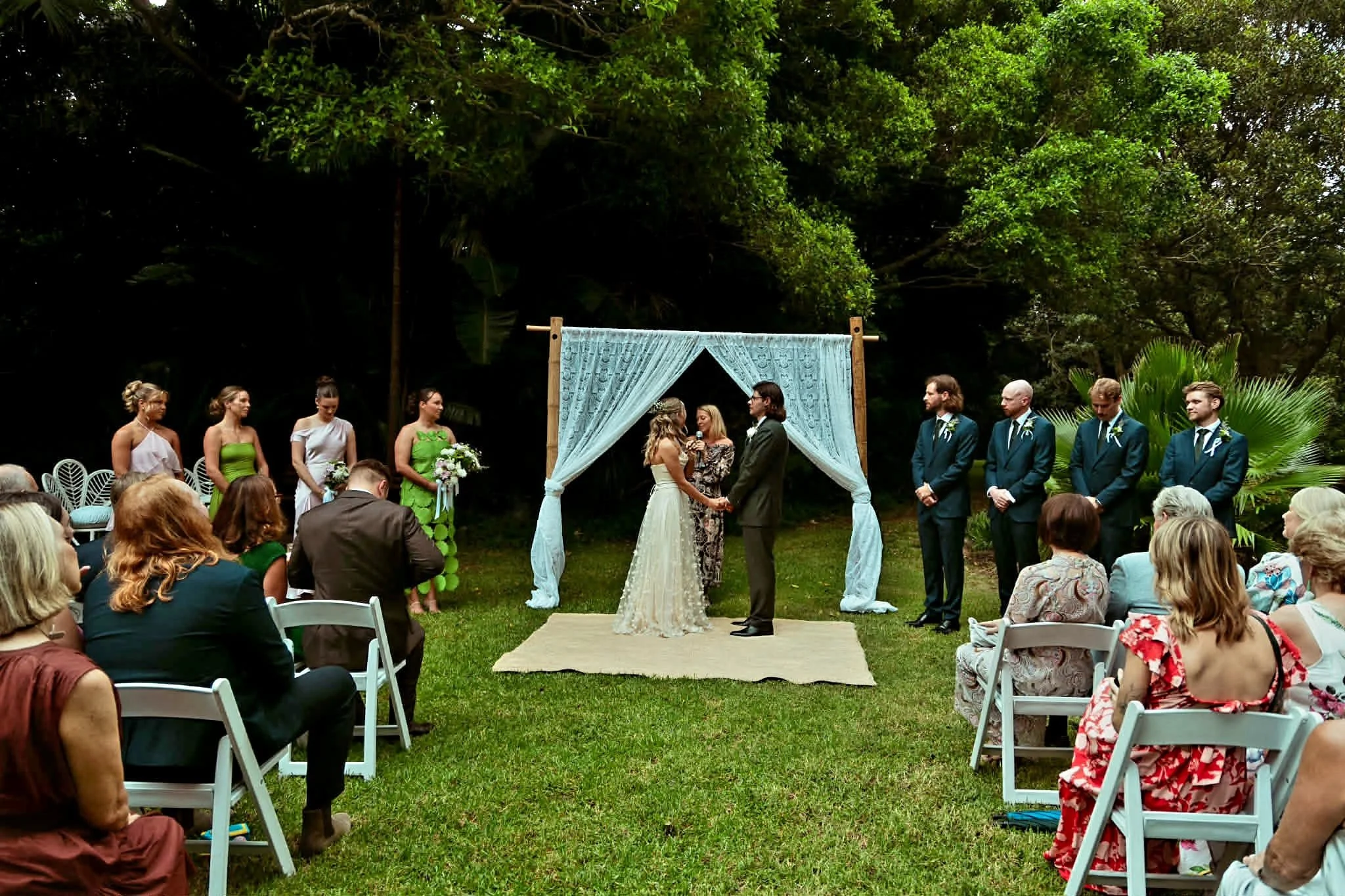 Bride and groom exchanging vows under an outdoor wedding arch, with bridesmaids and groomsmen standing nearby, guests seated in white chairs, surrounded by lush green trees.